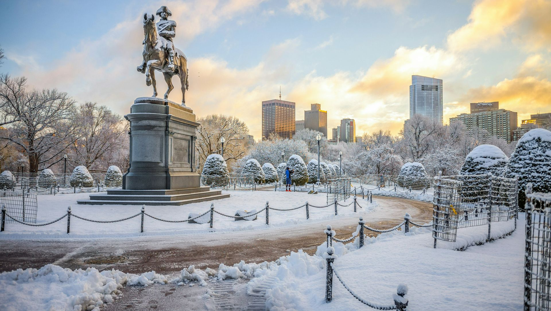 A statue of a rider on horseback in a snowy park, with a city skyline visible under a golden sunrise sky.