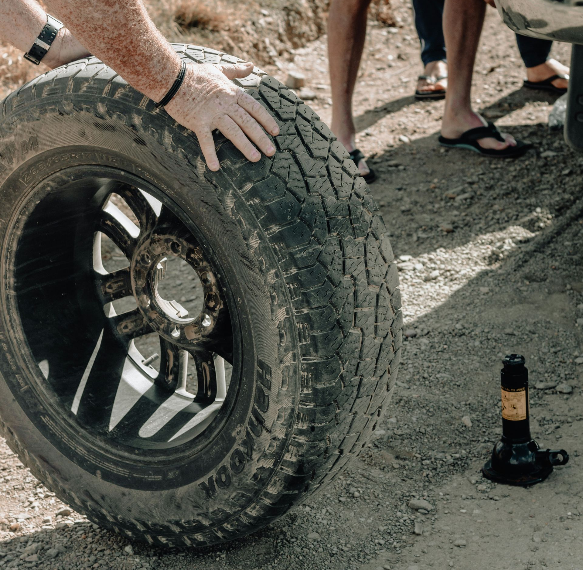 Two people changing a vehicle tire on a dirt road, with a jack positioned on the ground nearby.
