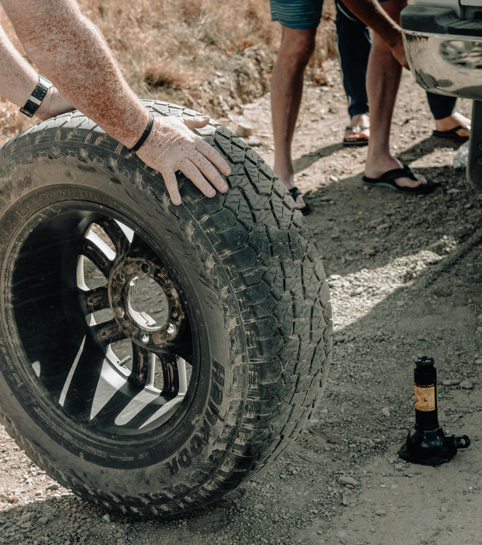 Two people changing a vehicle tire on a dirt road, with a jack positioned on the ground nearby.