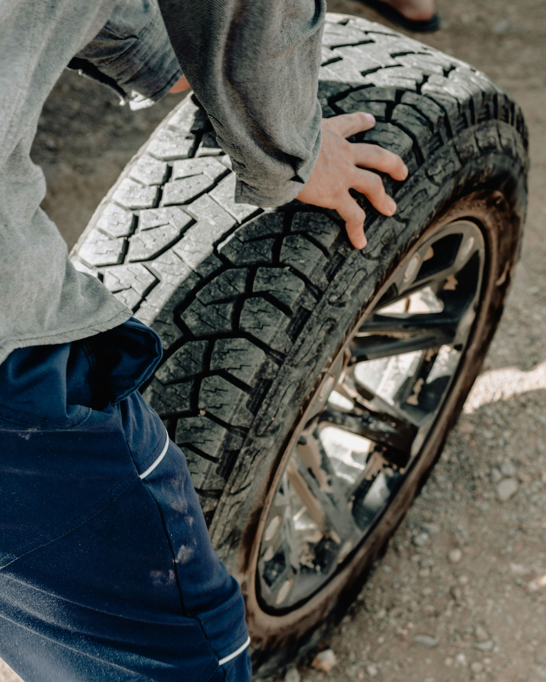 A person wearing a grey shirt and blue pants grips a large, treaded tire on a metal rim on a dirt surface.
