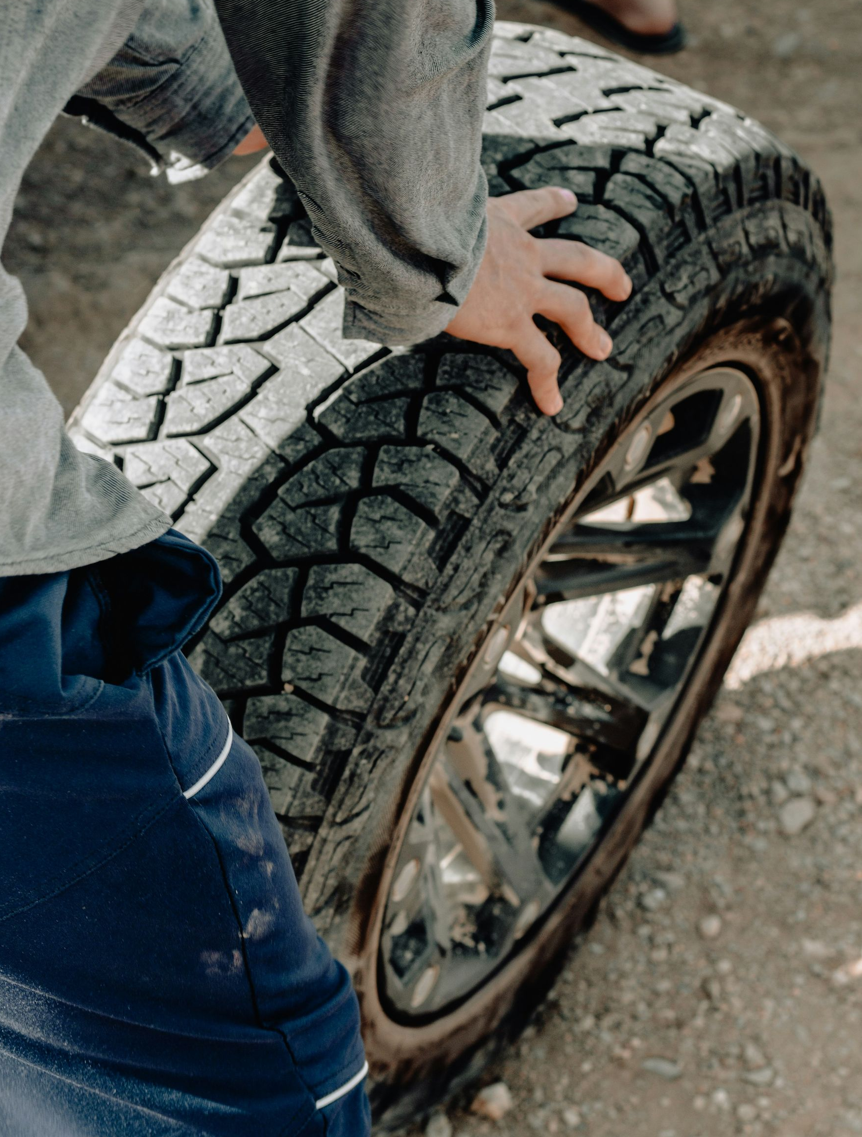 A person wearing a grey shirt and blue pants grips a large, treaded tire on a metal rim on a dirt surface.