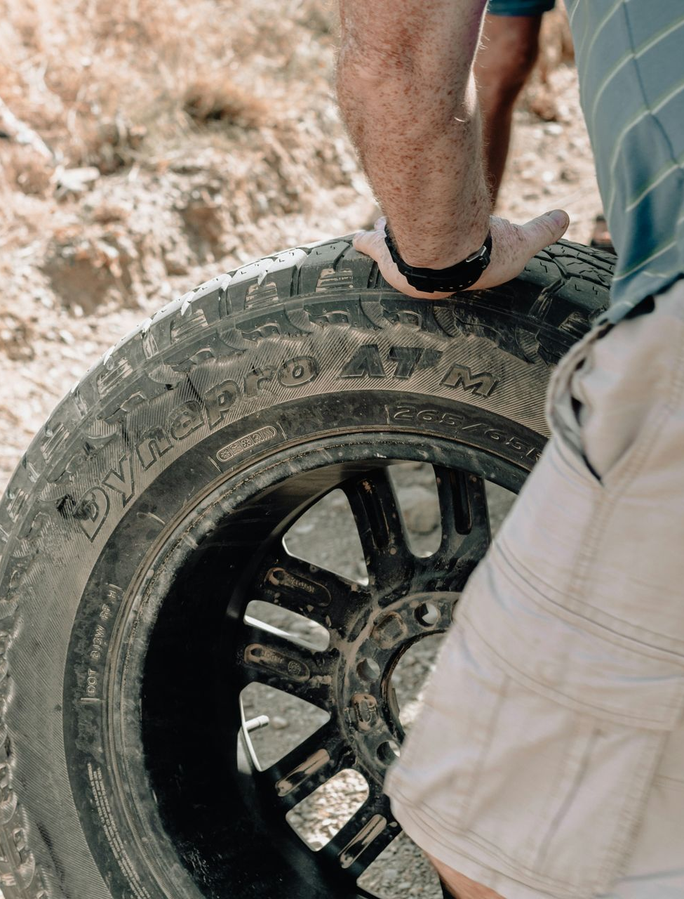Person mounting a black tire on a vehicle wheel in a dusty outdoor setting