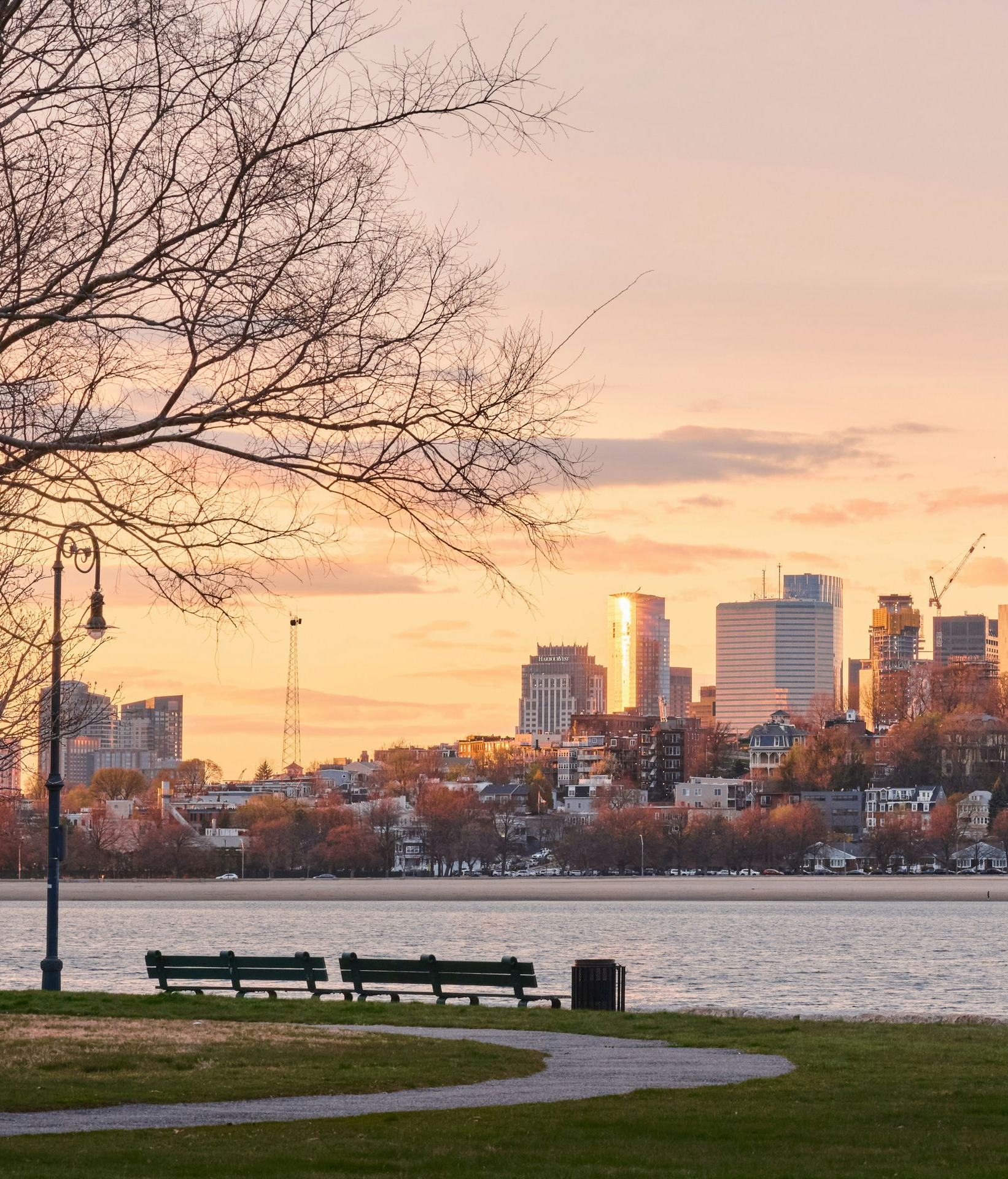 A sunset illuminates a city skyline across a calm body of water, viewed from a park with benches and a lamppost.