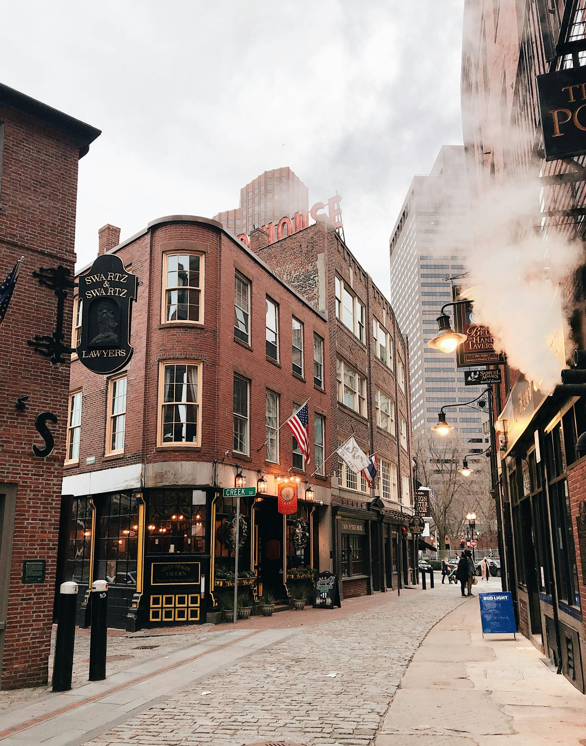 A historic brick corner building and cobblestone street in Boston, with steam rising near a dark iron pub sign.