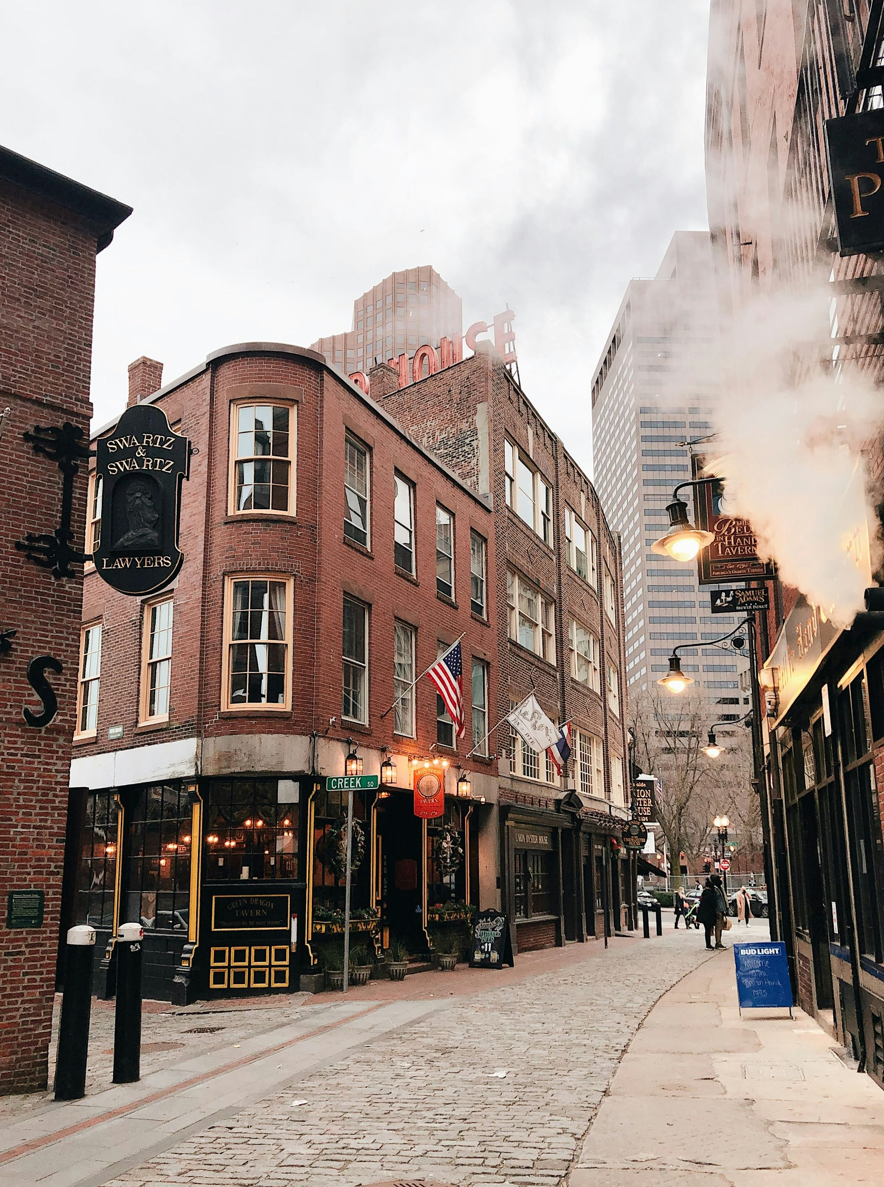 A historic brick corner building and cobblestone street in Boston, with steam rising near a dark iron pub sign.