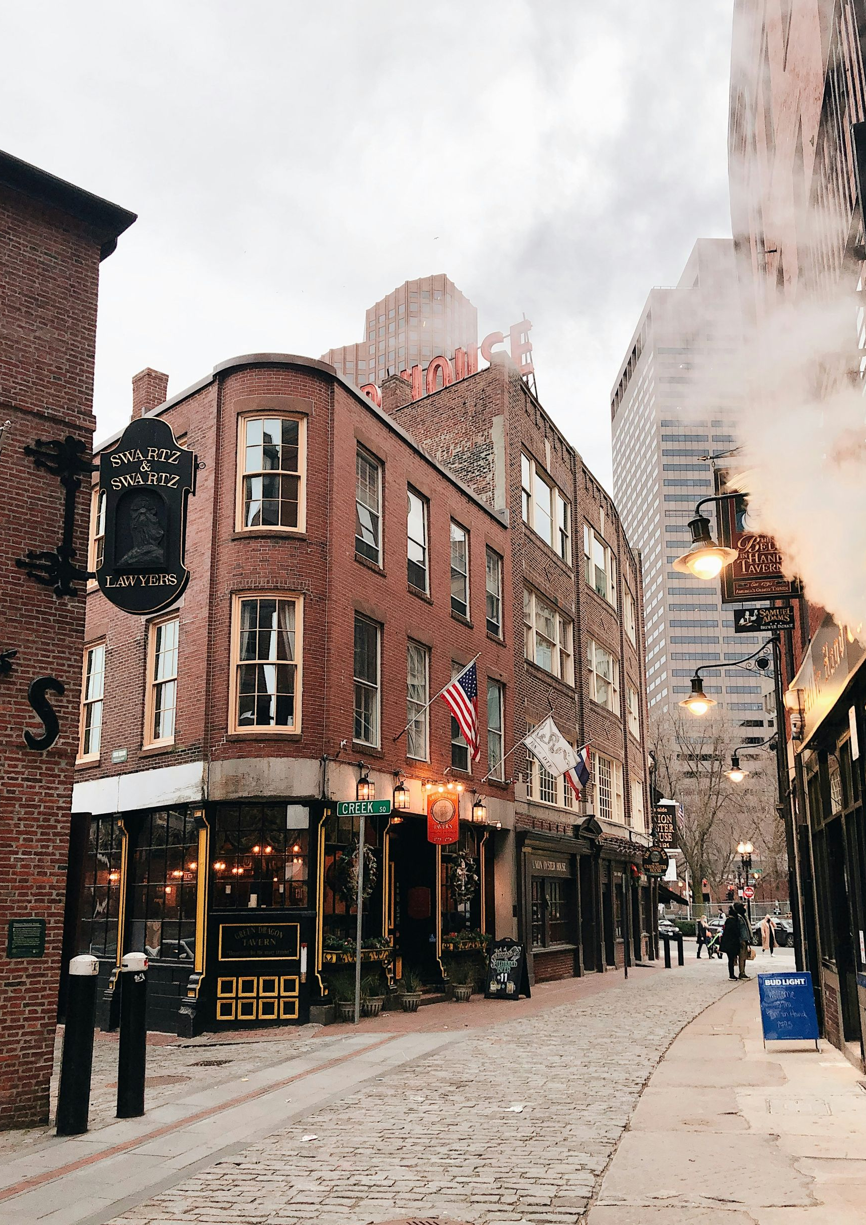 A historic brick corner building and cobblestone street in Boston, with steam rising near a dark iron pub sign.
