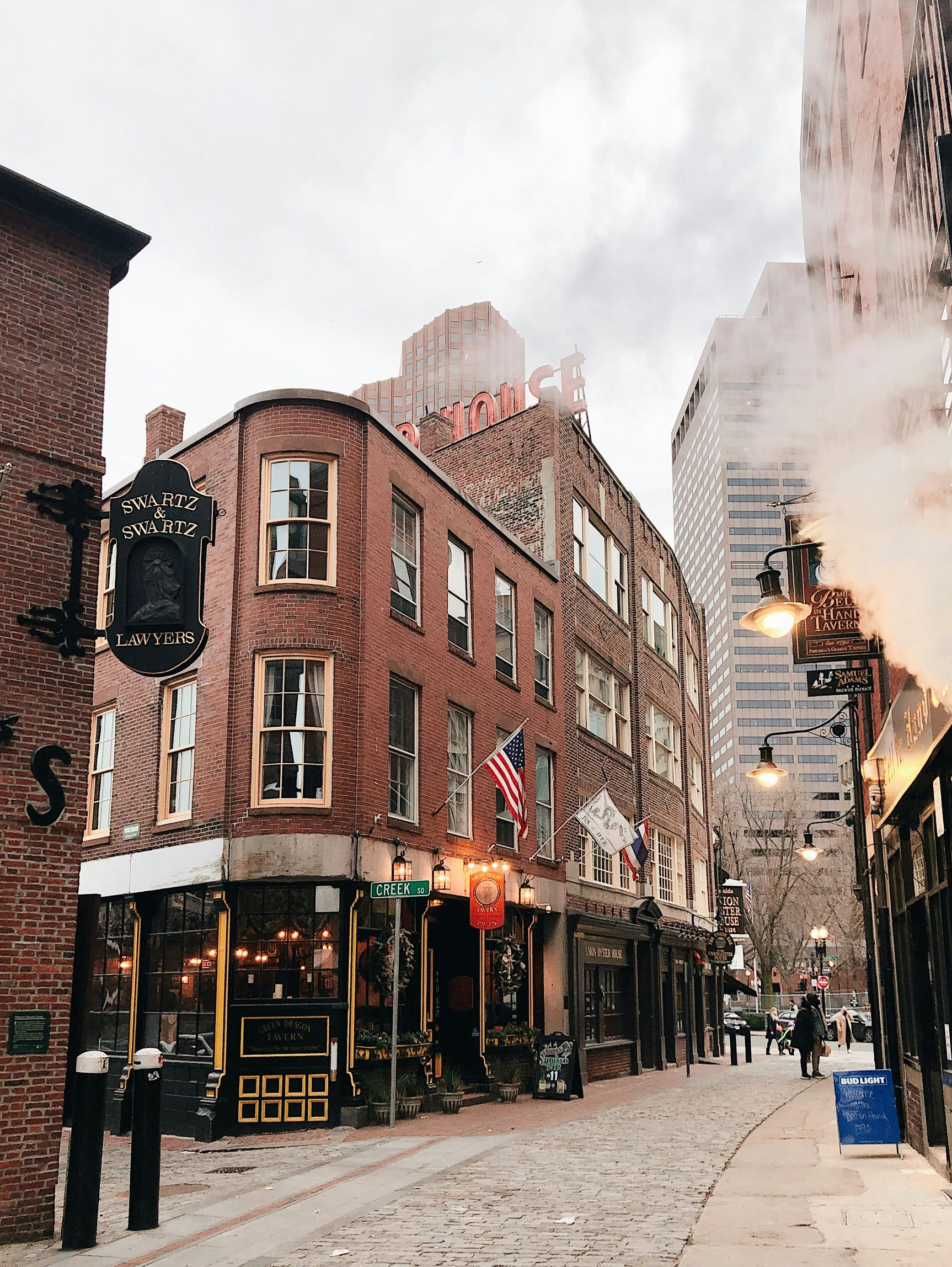 A historic brick corner building and cobblestone street in Boston, with steam rising near a dark iron pub sign.