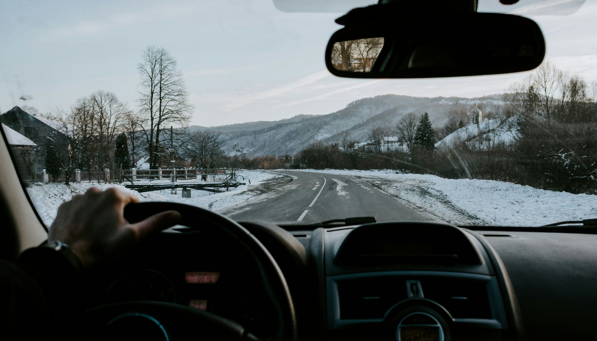 Driver's perspective of a snowy road winding through a winter landscape, with mountains in the background.