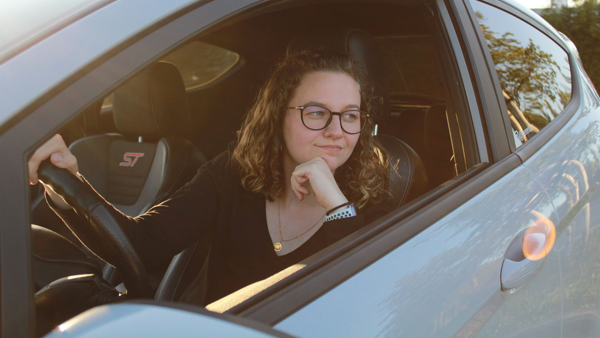 Person with glasses sitting in a car, resting chin on hand and looking out the open window.