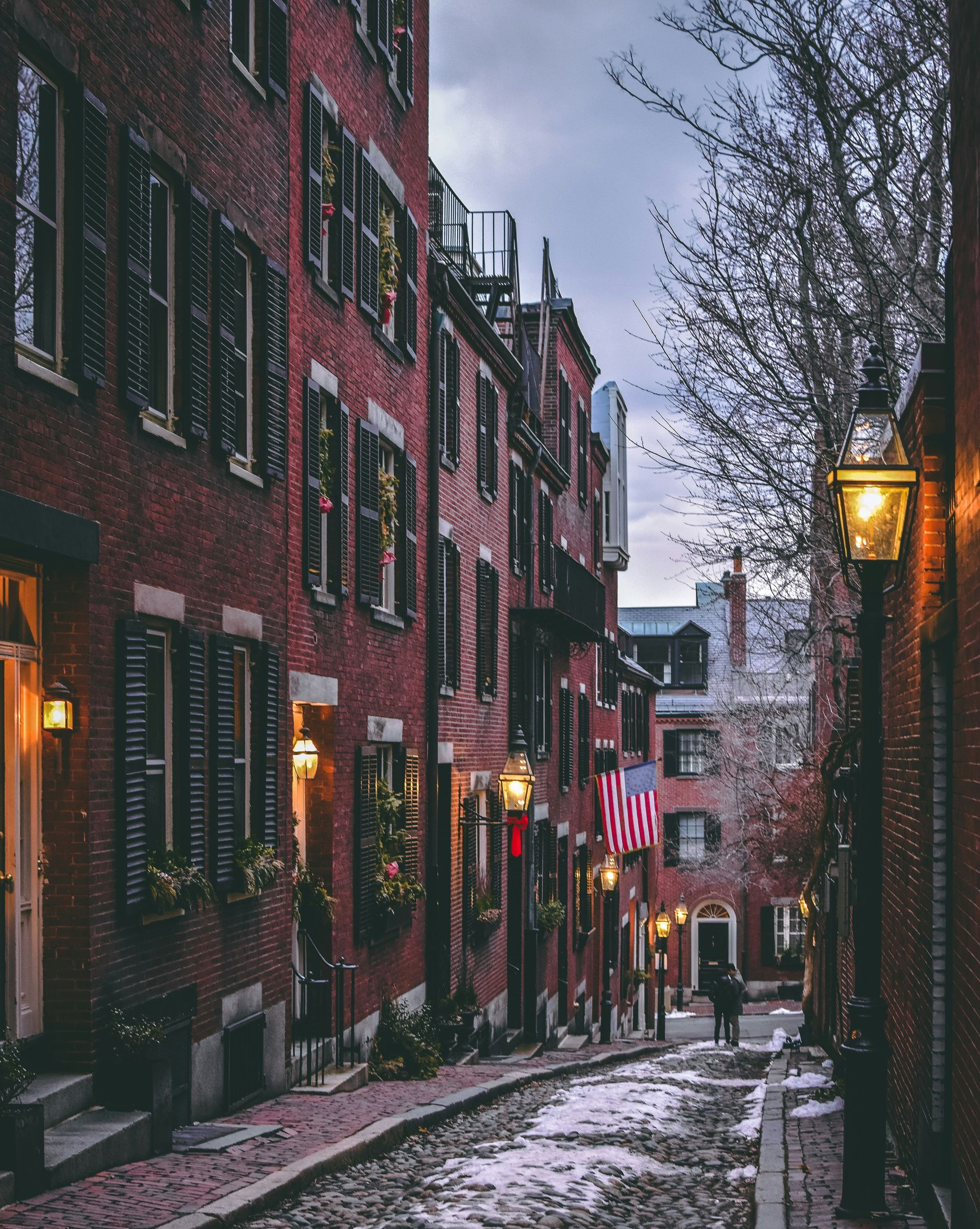 A cobblestone street slopes between historic brick buildings with dark shutters, illuminated by glowing streetlamps.