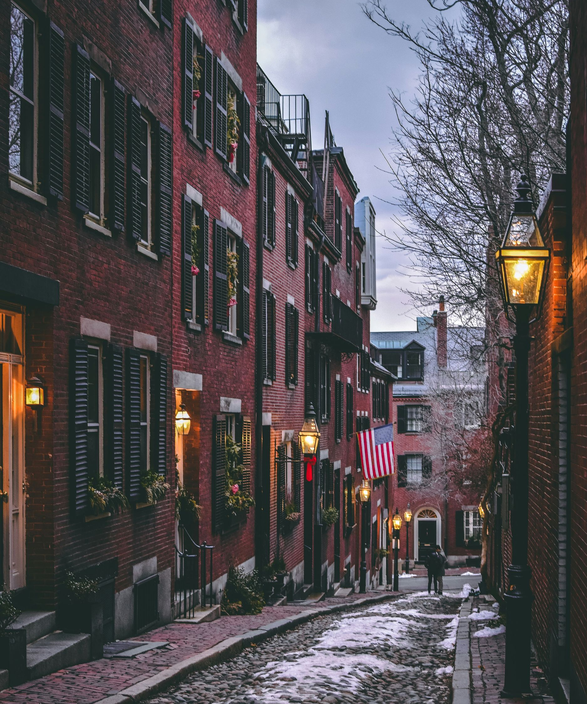 A cobblestone street slopes between historic brick buildings with dark shutters, illuminated by glowing streetlamps.