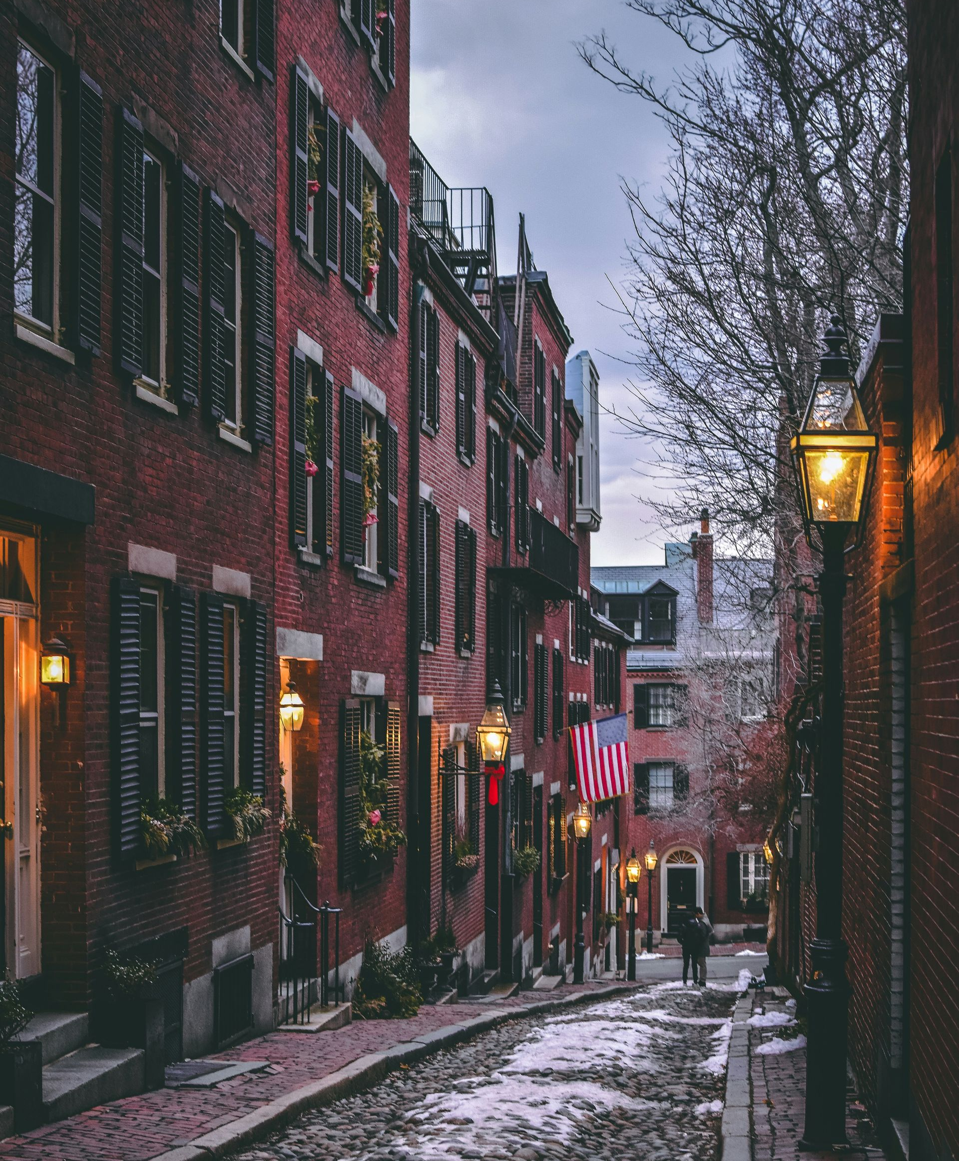 A cobblestone street slopes between historic brick buildings with dark shutters, illuminated by glowing streetlamps.