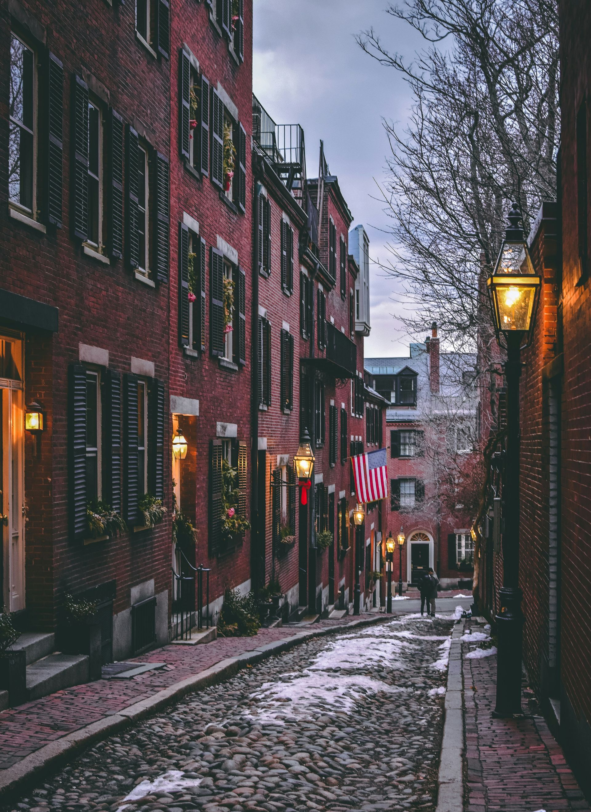 A cobblestone street slopes between historic brick buildings with dark shutters, illuminated by glowing streetlamps.