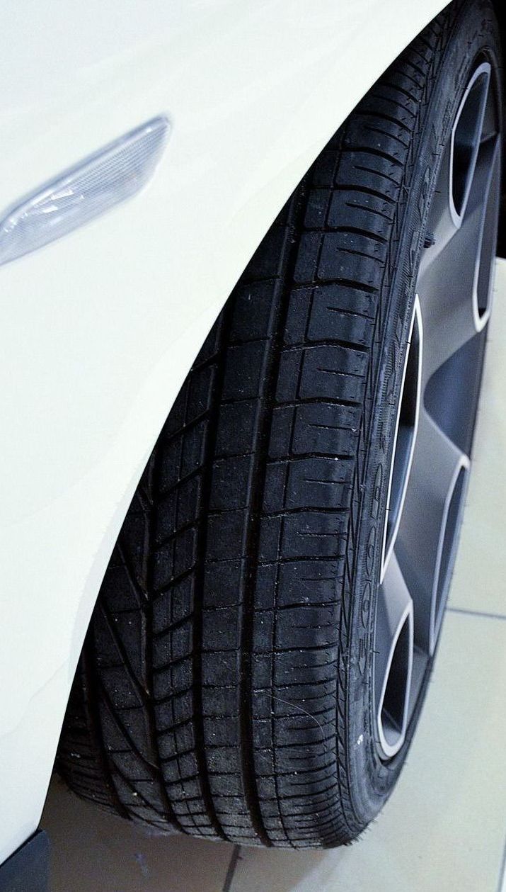 White car's front tire and alloy wheel close-up. The black tire has tread, and the wheel has a silver rim.