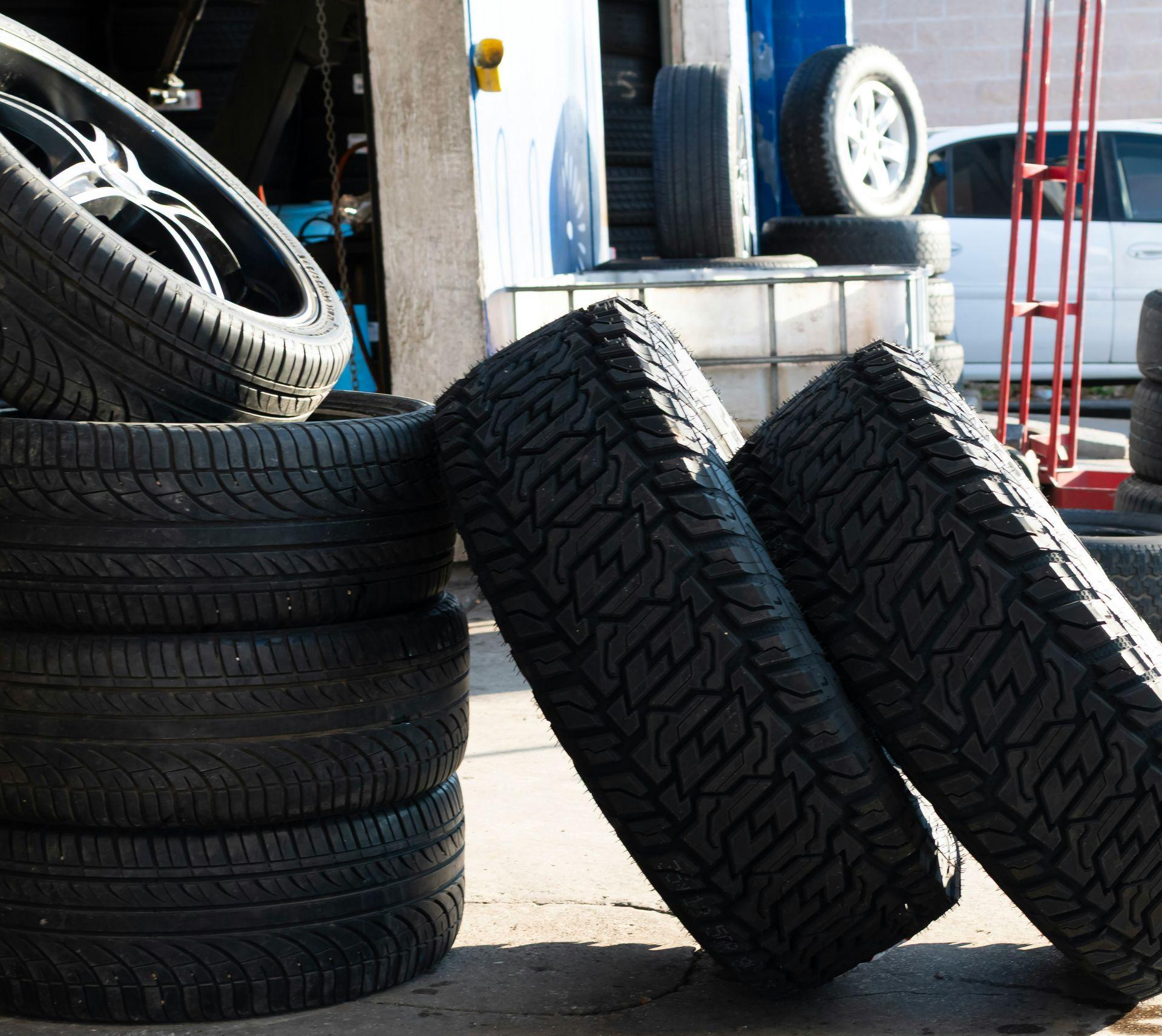 Stacked tires outside a garage, with two leaning against a curb on a sunny pavement.