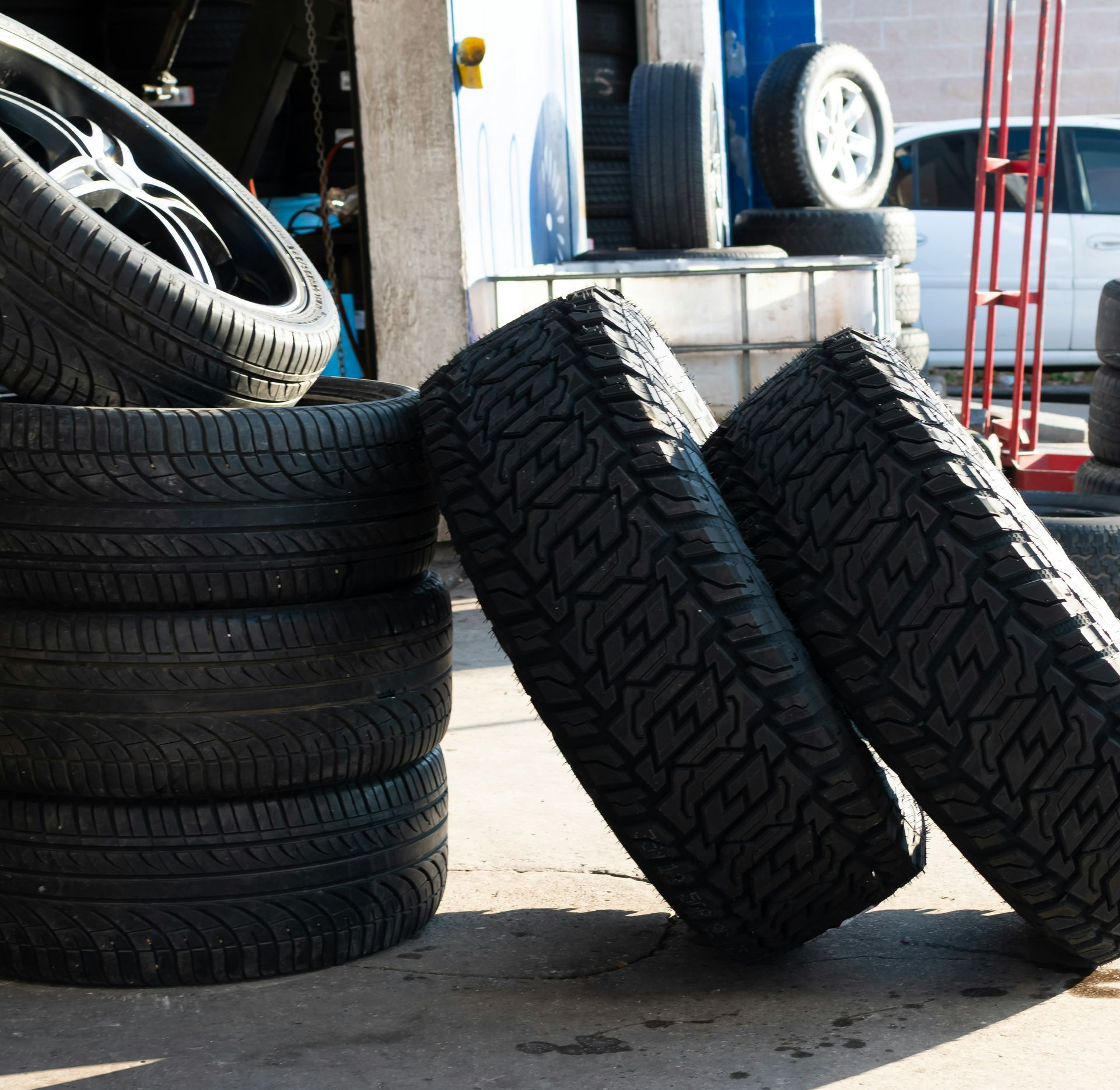 Stacked tires outside a garage, with two leaning against a curb on a sunny pavement.