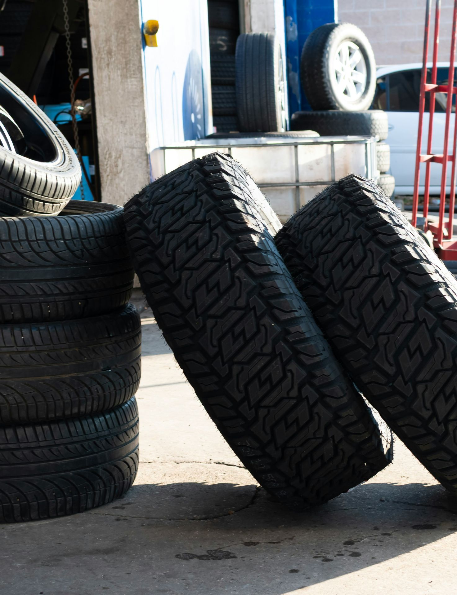 Stacked tires outside a garage, with two leaning against a curb on a sunny pavement.