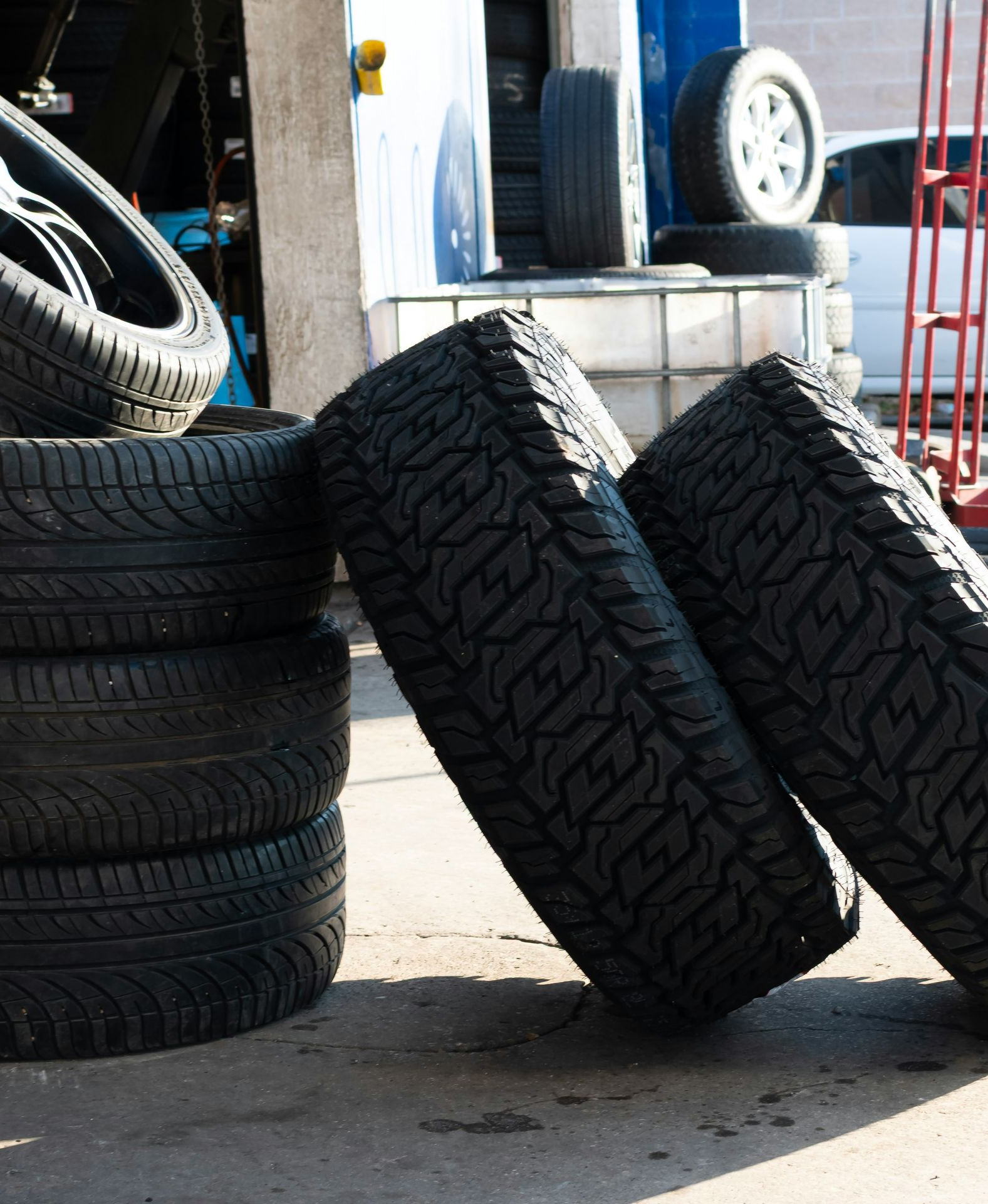Stacked tires outside a garage, with two leaning against a curb on a sunny pavement.