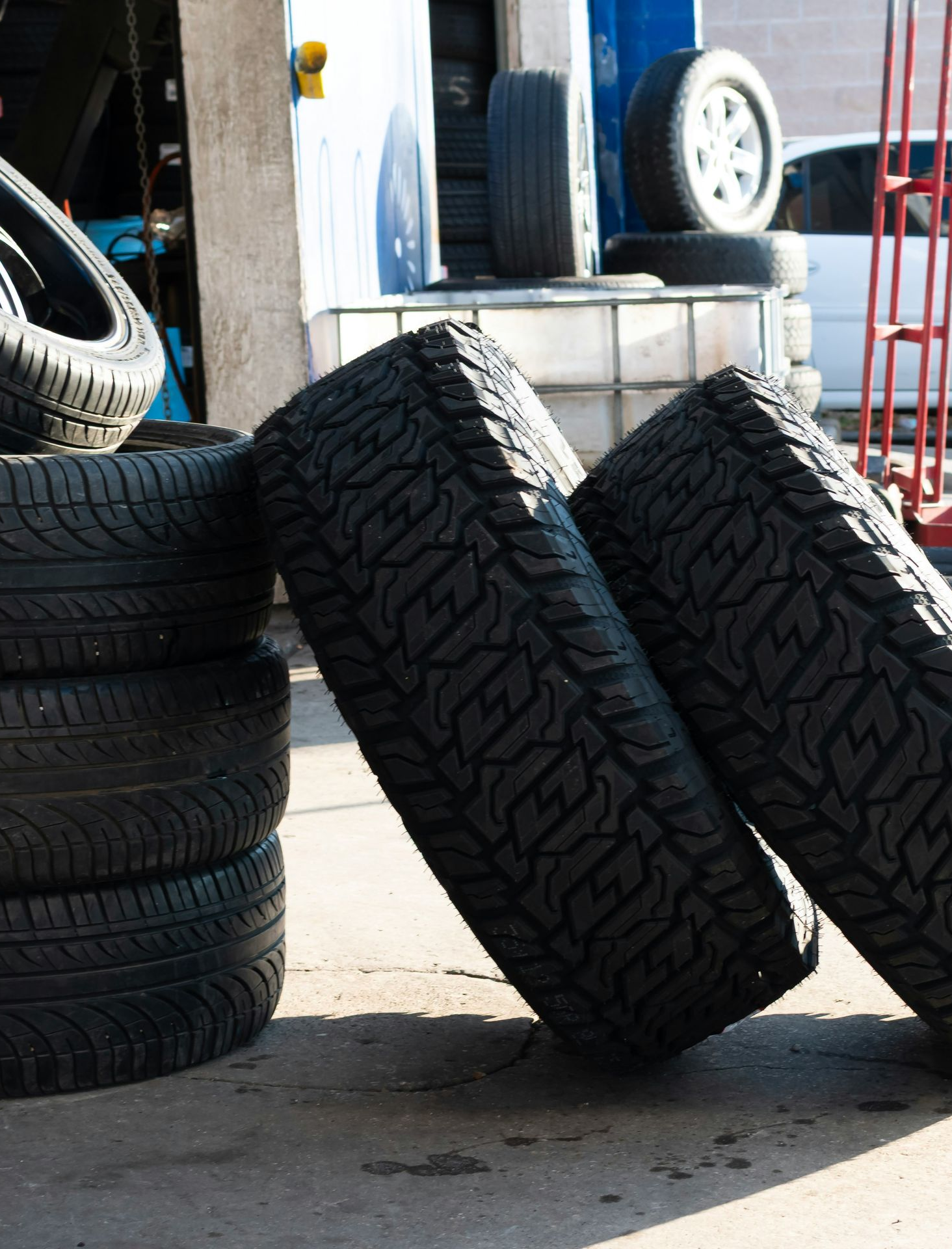 Stacked tires outside a garage, with two leaning against a curb on a sunny pavement.