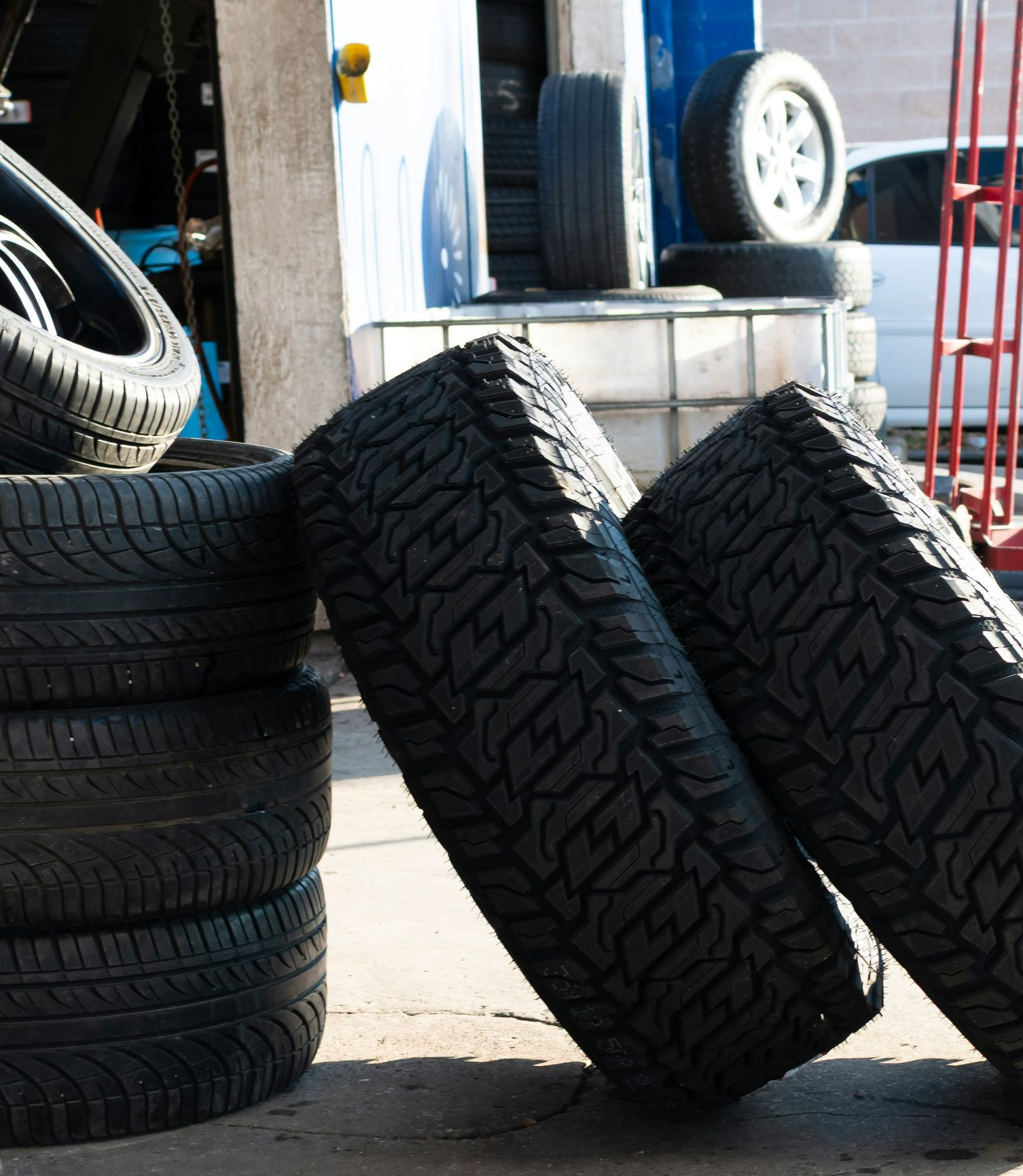 Stacked tires outside a garage, with two leaning against a curb on a sunny pavement.