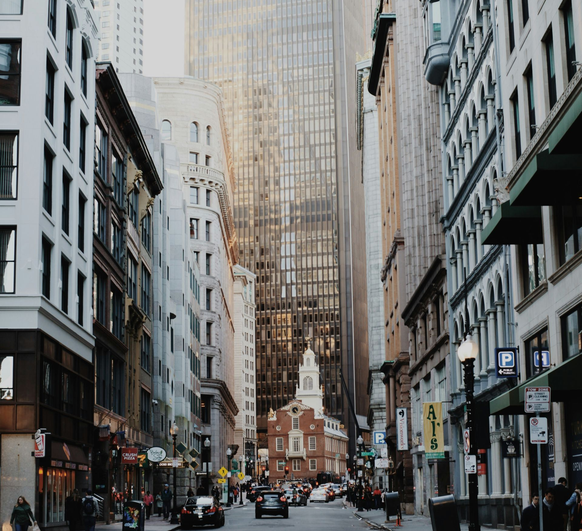 A narrow city street lined with tall buildings leads toward an old brick building at the end, under a hazy sky.