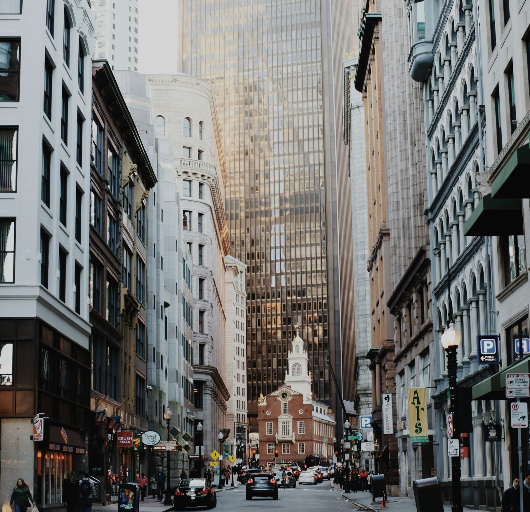 A narrow city street lined with tall buildings leads toward an old brick building at the end, under a hazy sky.