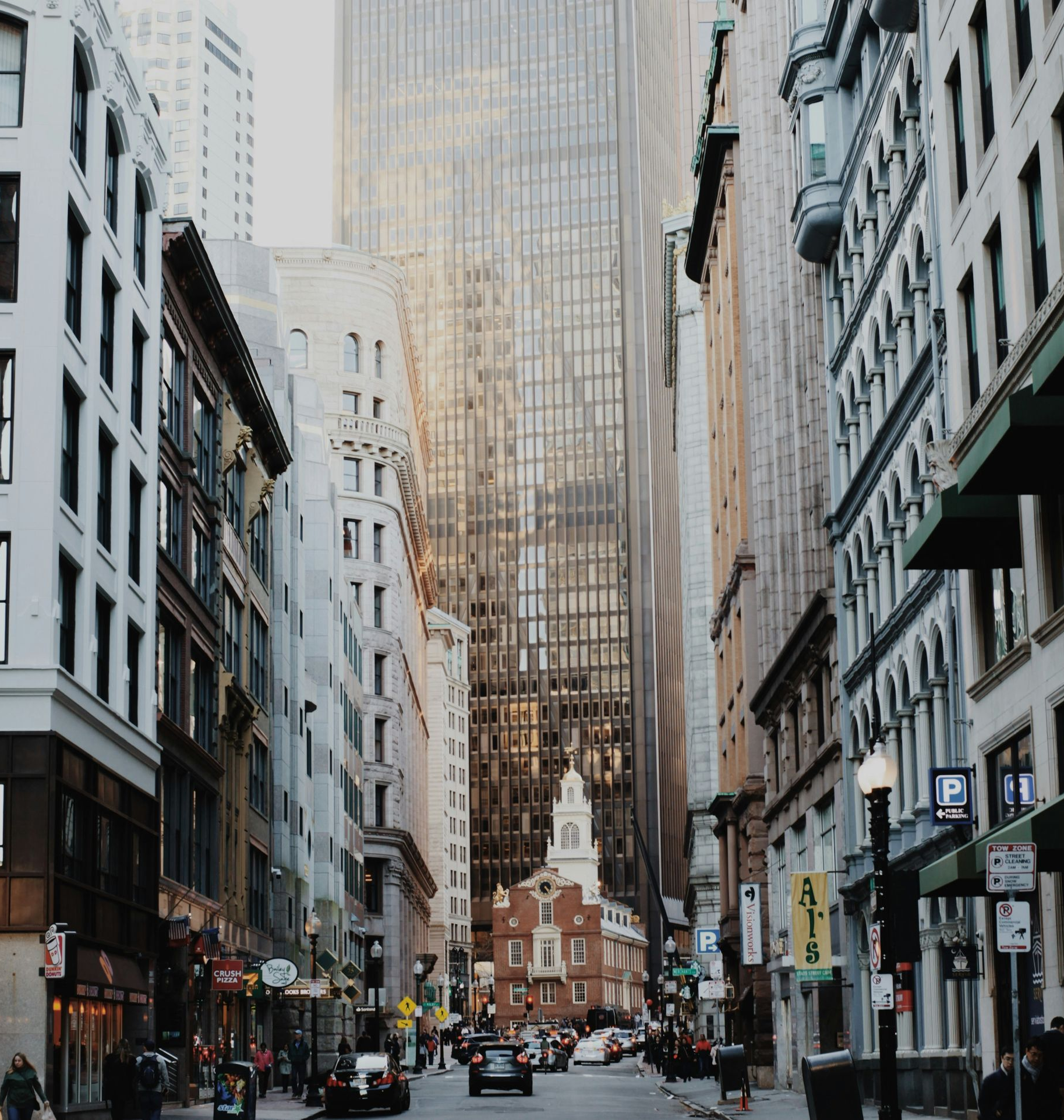 A narrow city street lined with tall buildings leads toward an old brick building at the end, under a hazy sky.