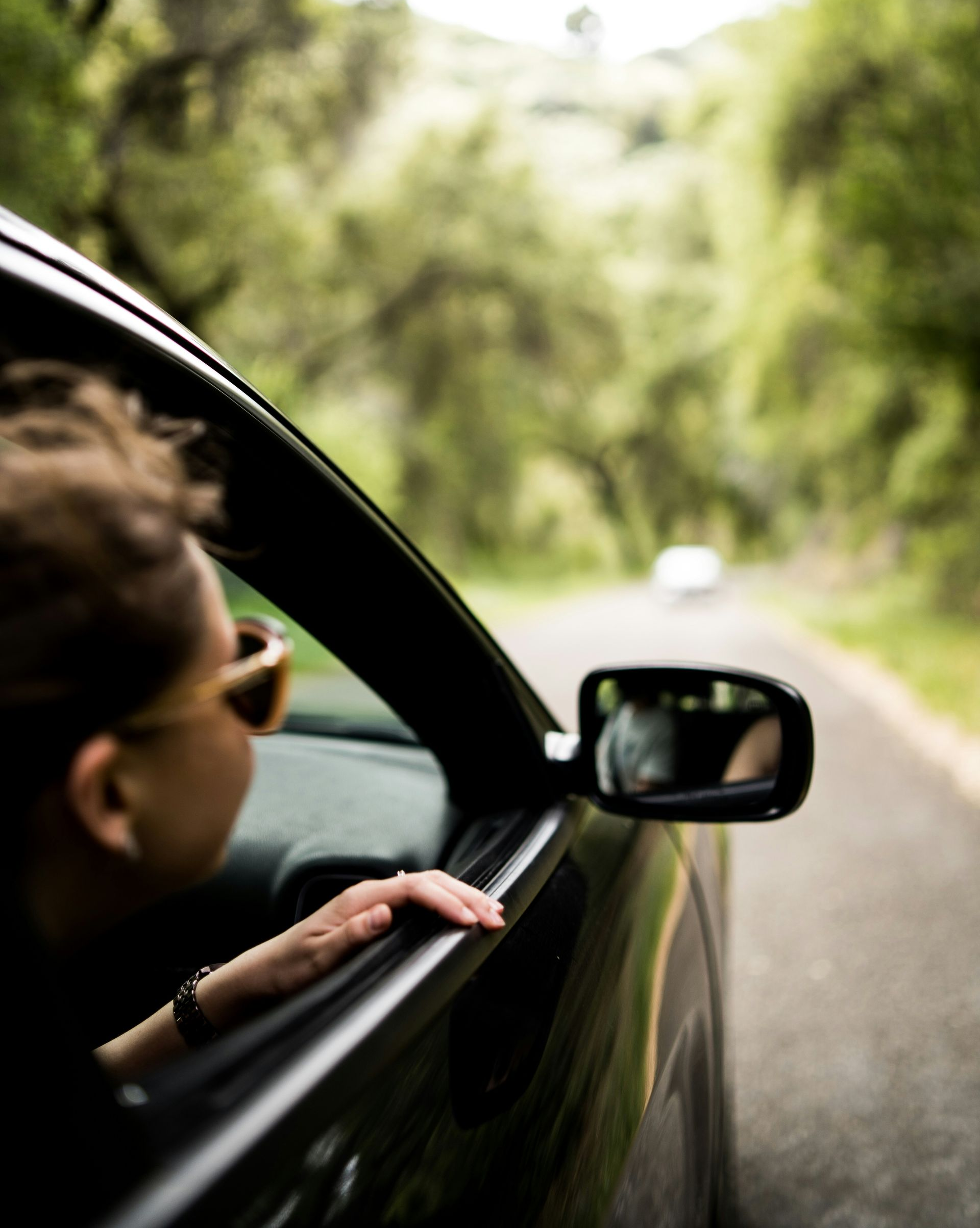 Person leaning out of a car window on a tree-lined road, with a side mirror in view.