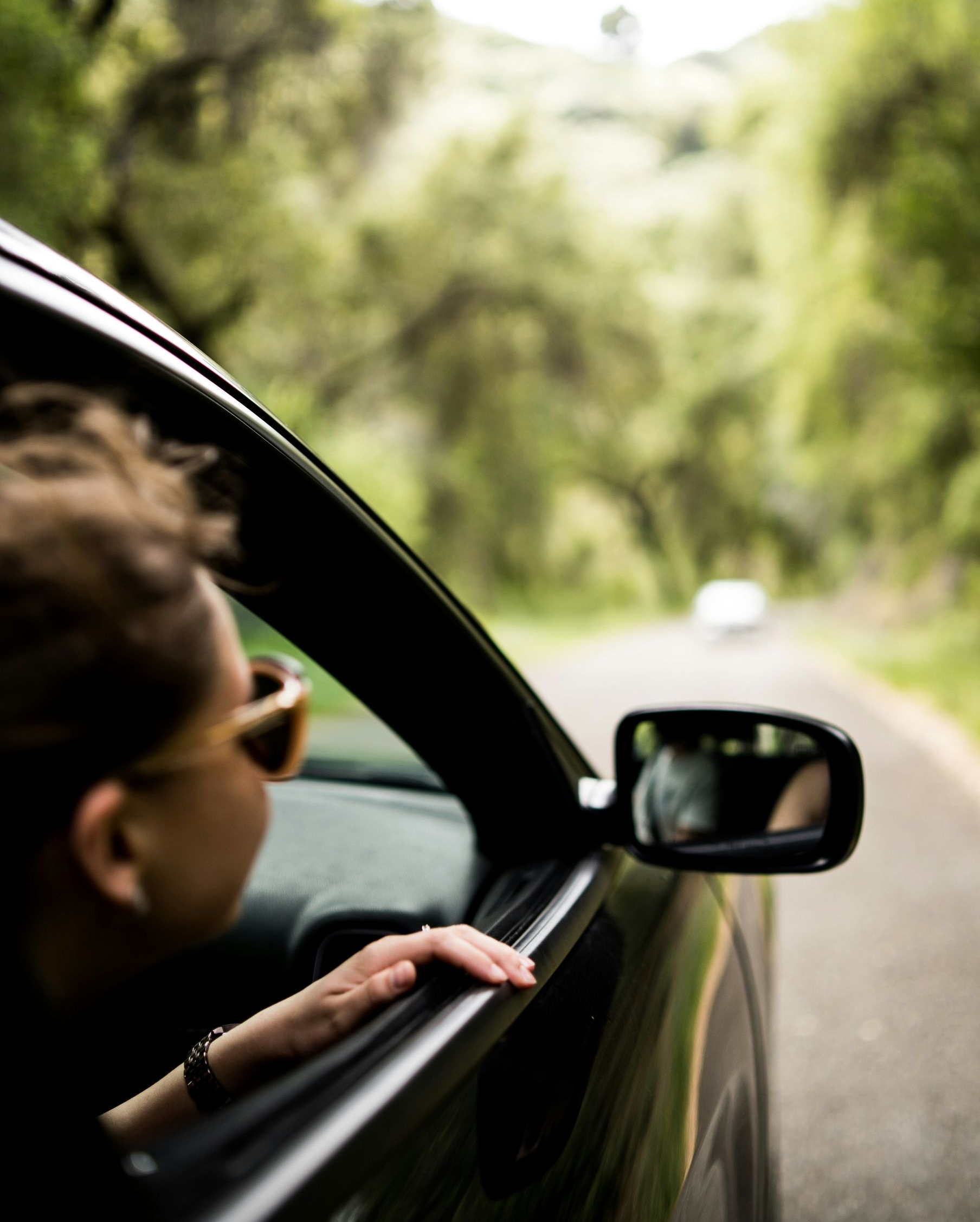 Person leaning out of a car window on a tree-lined road, with a side mirror in view.