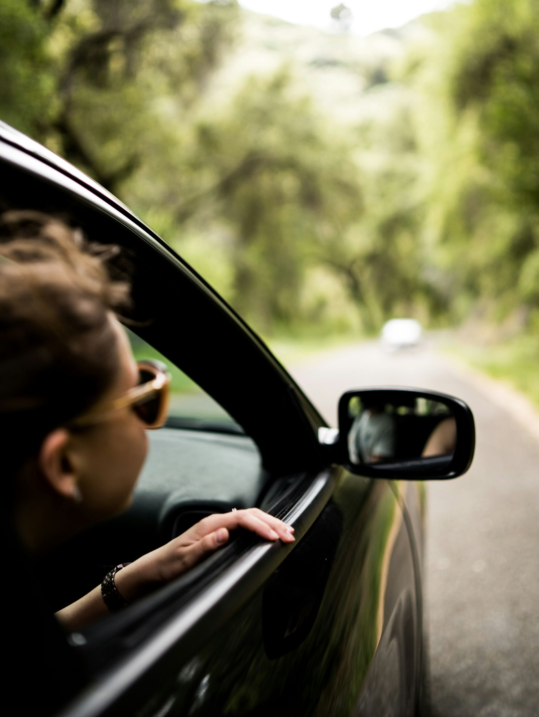 Person leaning out of a car window on a tree-lined road, with a side mirror in view.