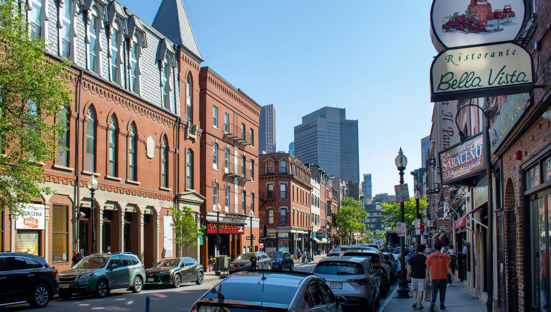 A sunny street scene in Boston's North End, featuring red brick buildings, a prominent Bella Vista sign, and parked cars.