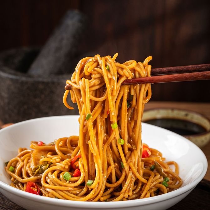 Chopsticks lifting saucy noodles from a white bowl, garnished with vegetables
