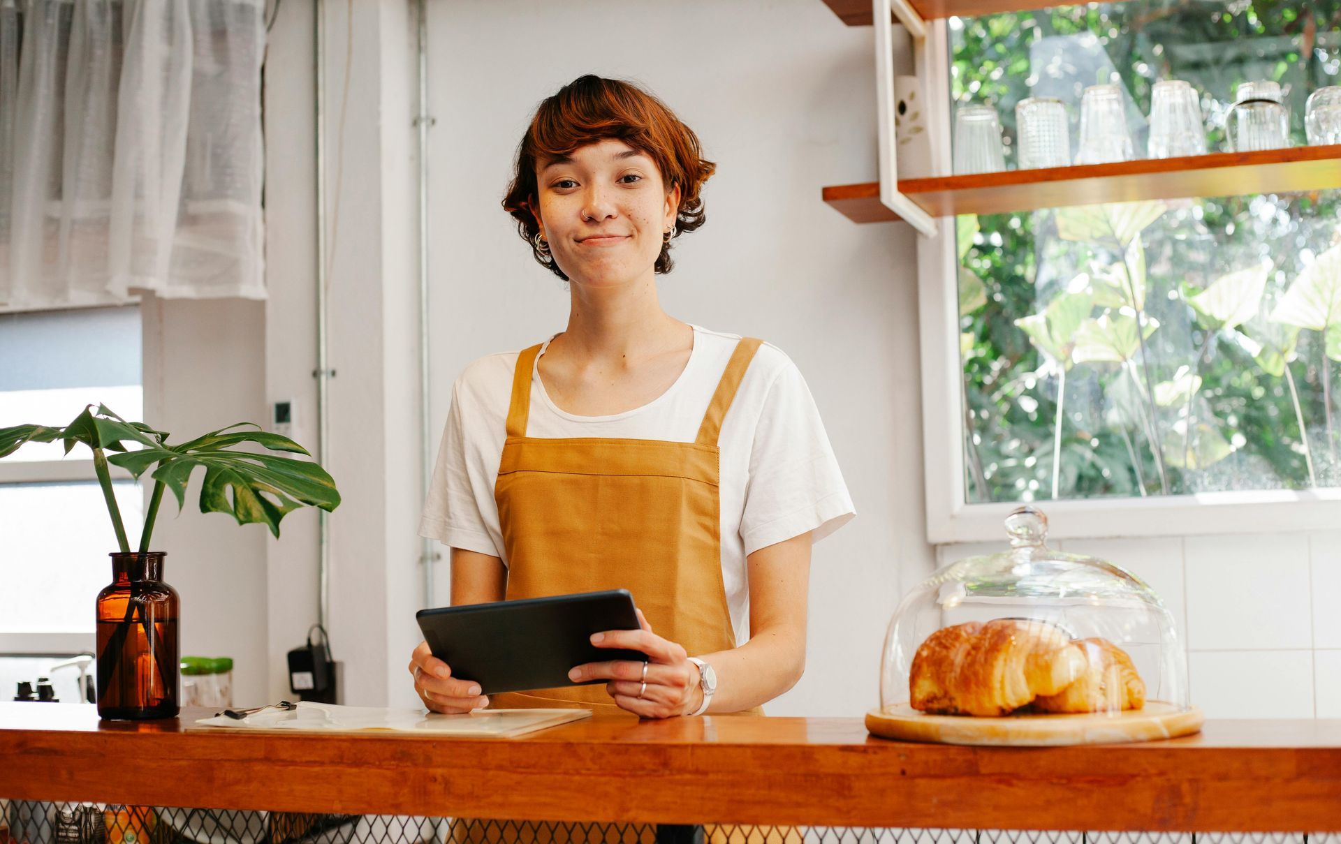 Person in a yellow apron smiles, holding a tablet behind a cafe counter with a croissant display and a window.