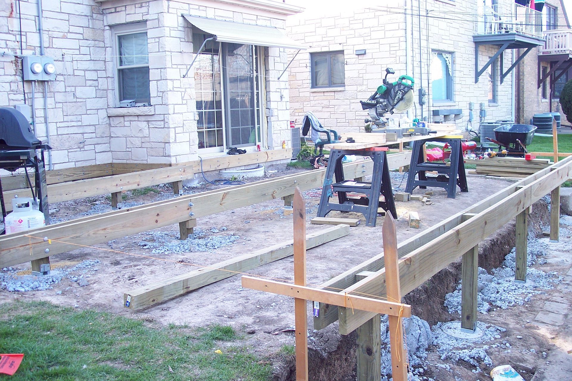 Construction site with wooden forms and tools beside a stone house, ground covered in snow.