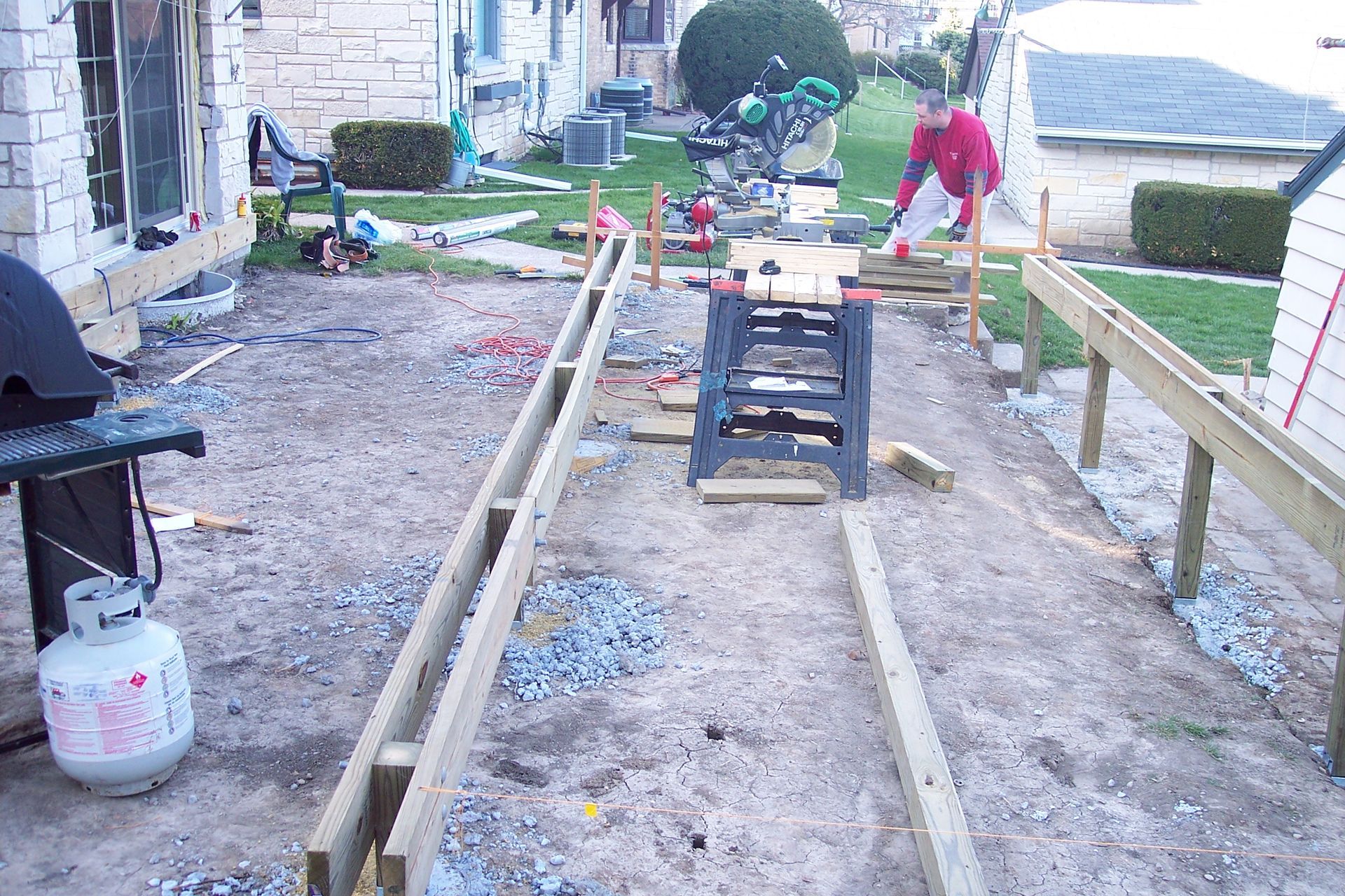 Two workers laying long wooden beams on a gravel driveway beside a house under construction.