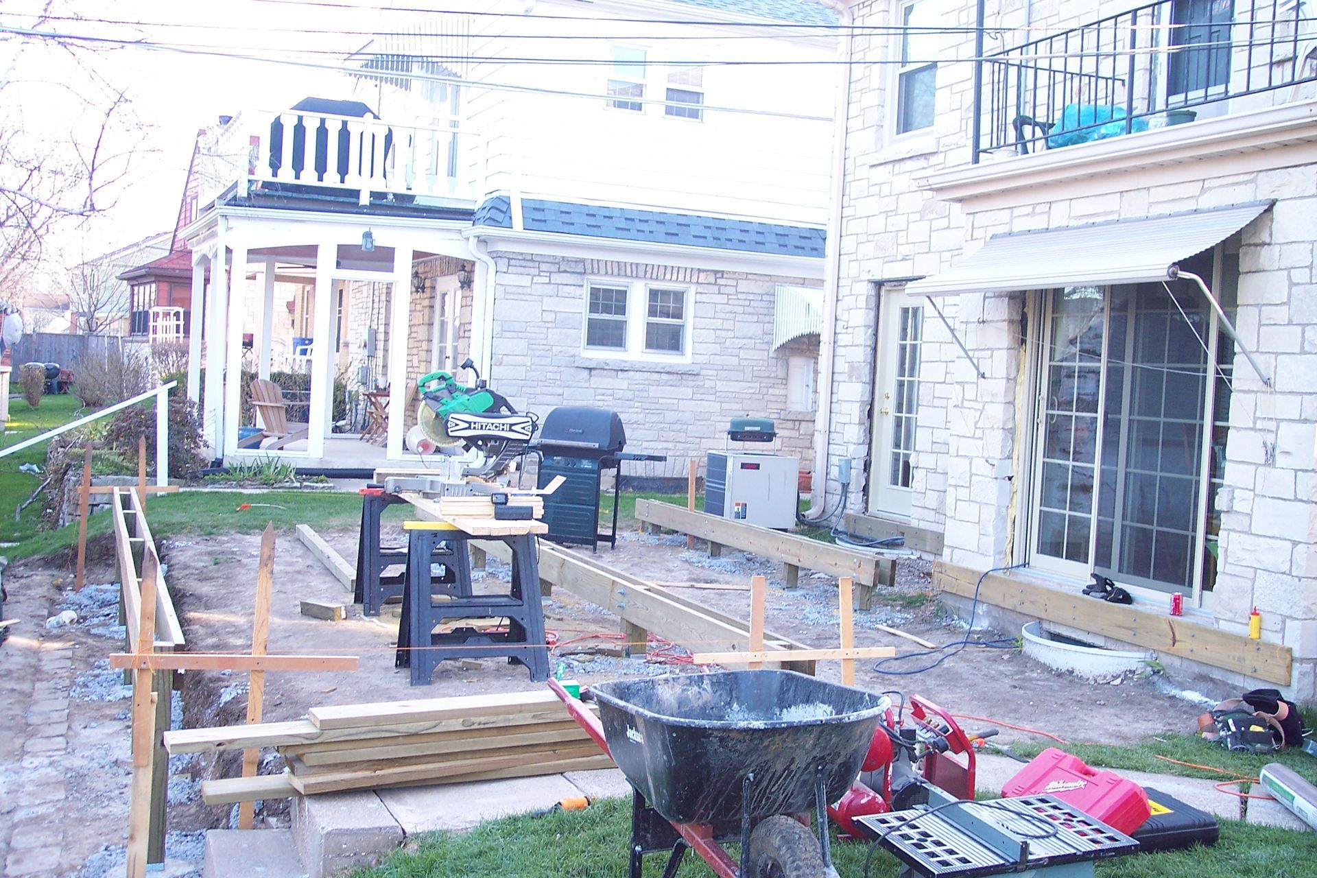 Backyard patio under construction with lumber, tools, a grill, and a stone house backdrop