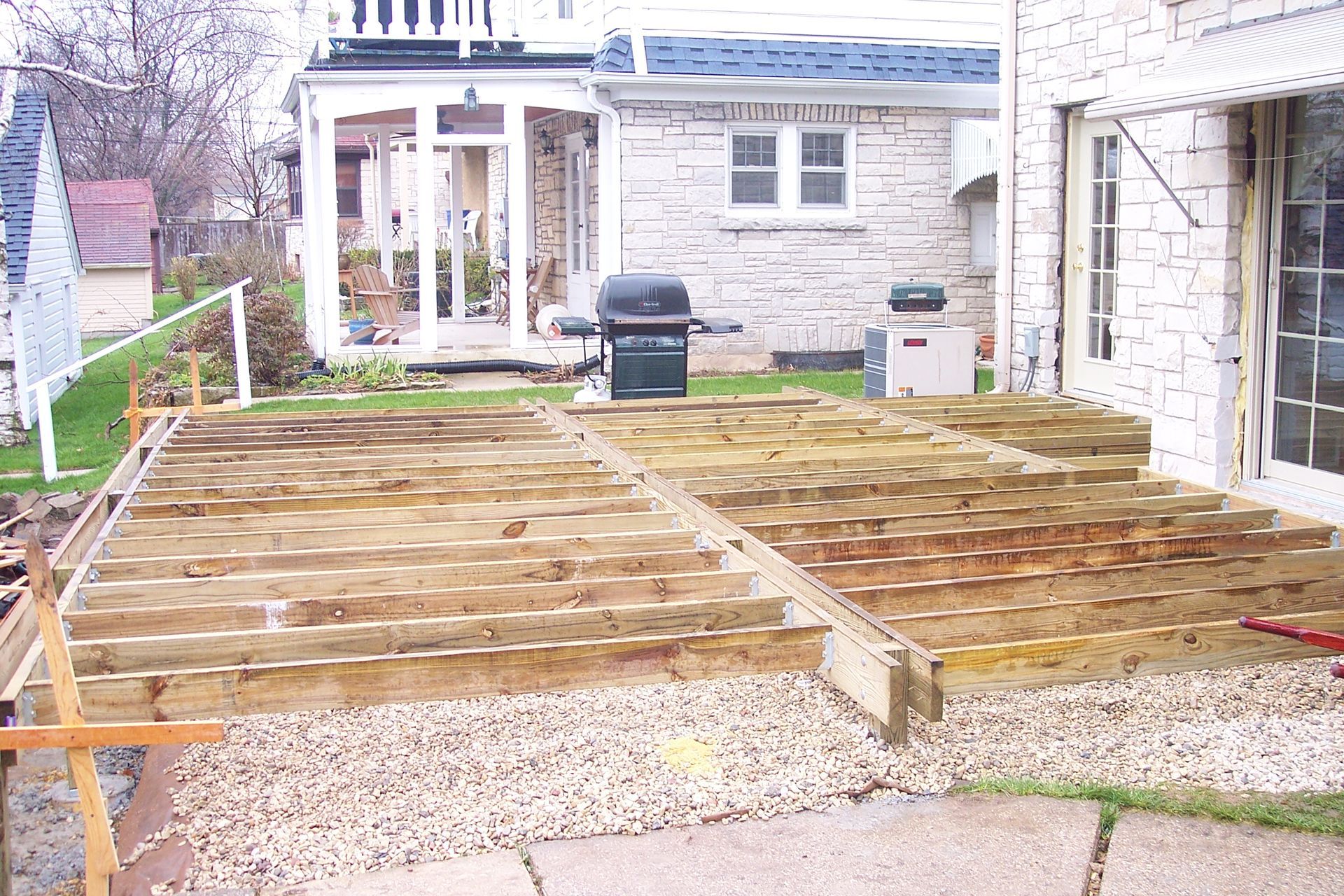 Wooden deck framework under construction behind a stone house, with gravel and a white railing nearby.