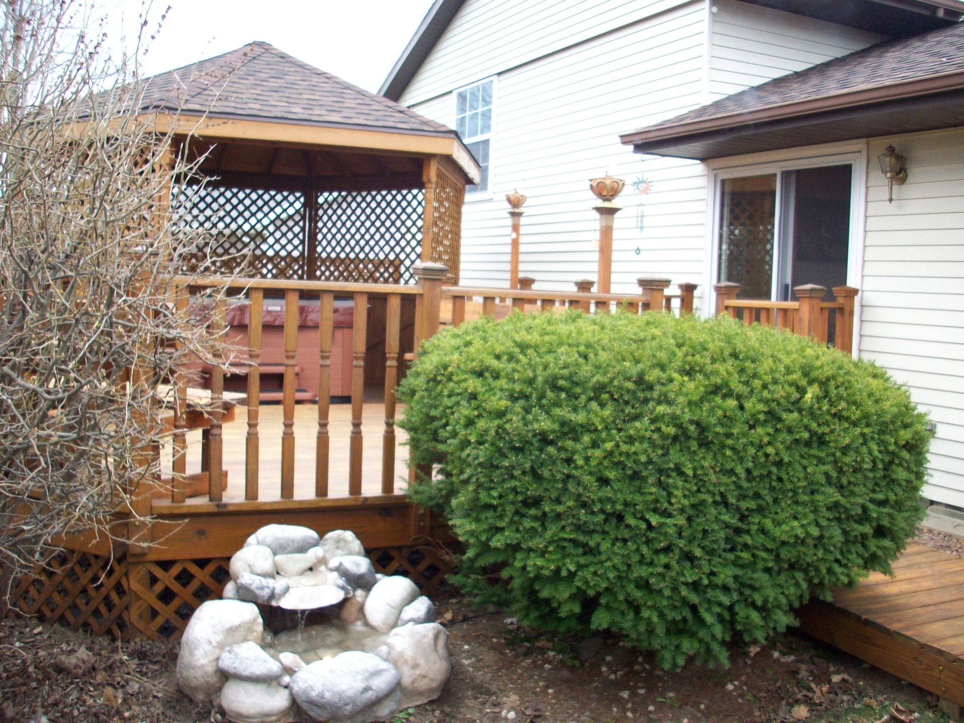 Backyard deck and gazebo with wooden railing, green shrub, and small stone fountain