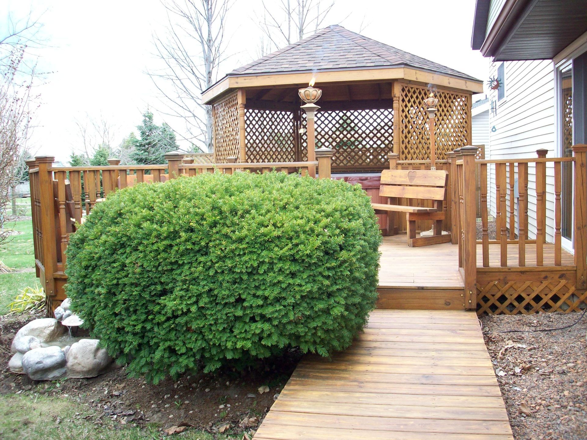 Wooden gazebo on a backyard deck with lattice sides, a large green shrub, and a walkway in front
