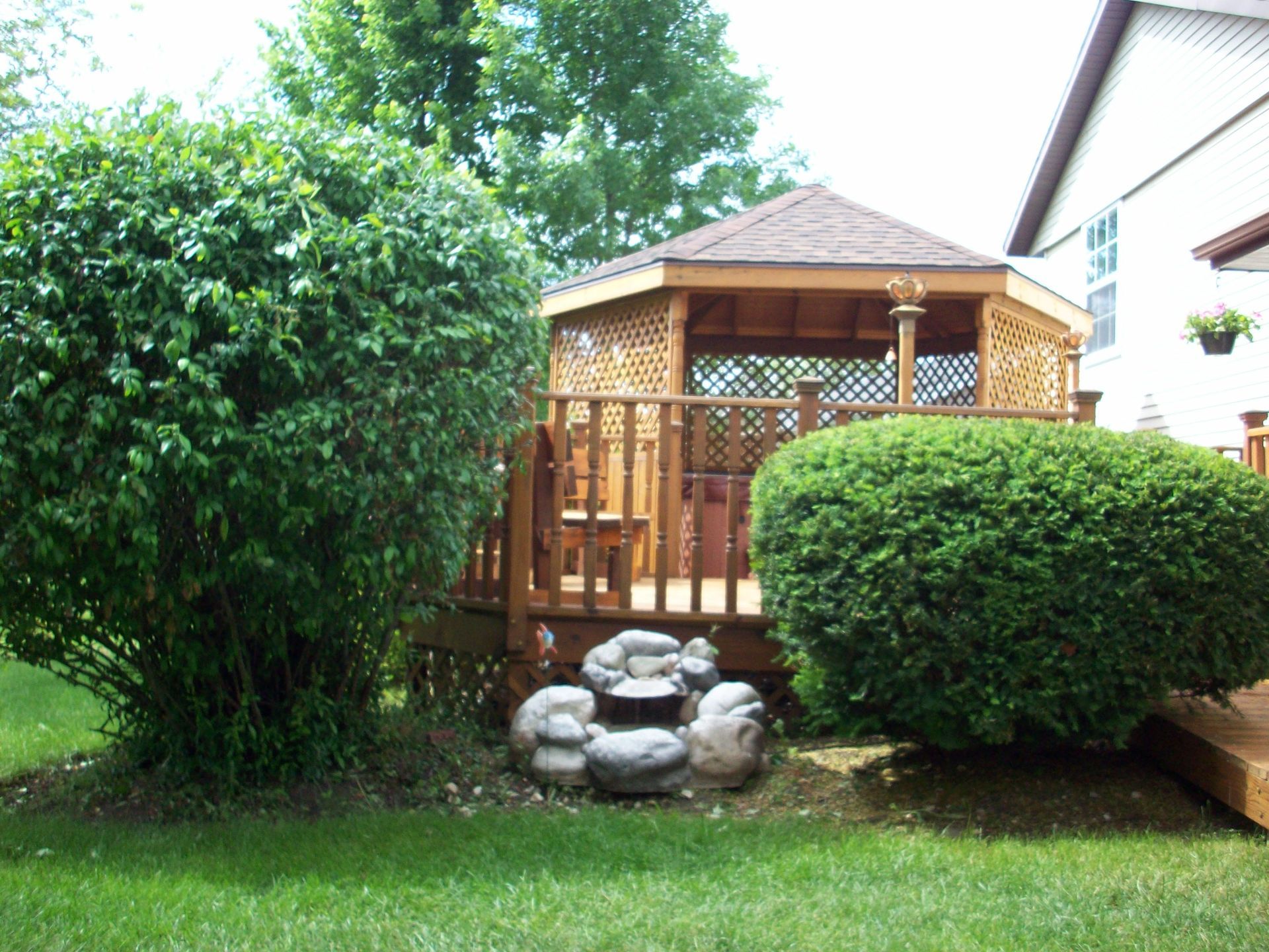 Backyard gazebo with wooden deck, trimmed shrubs, and a stone fountain under trees