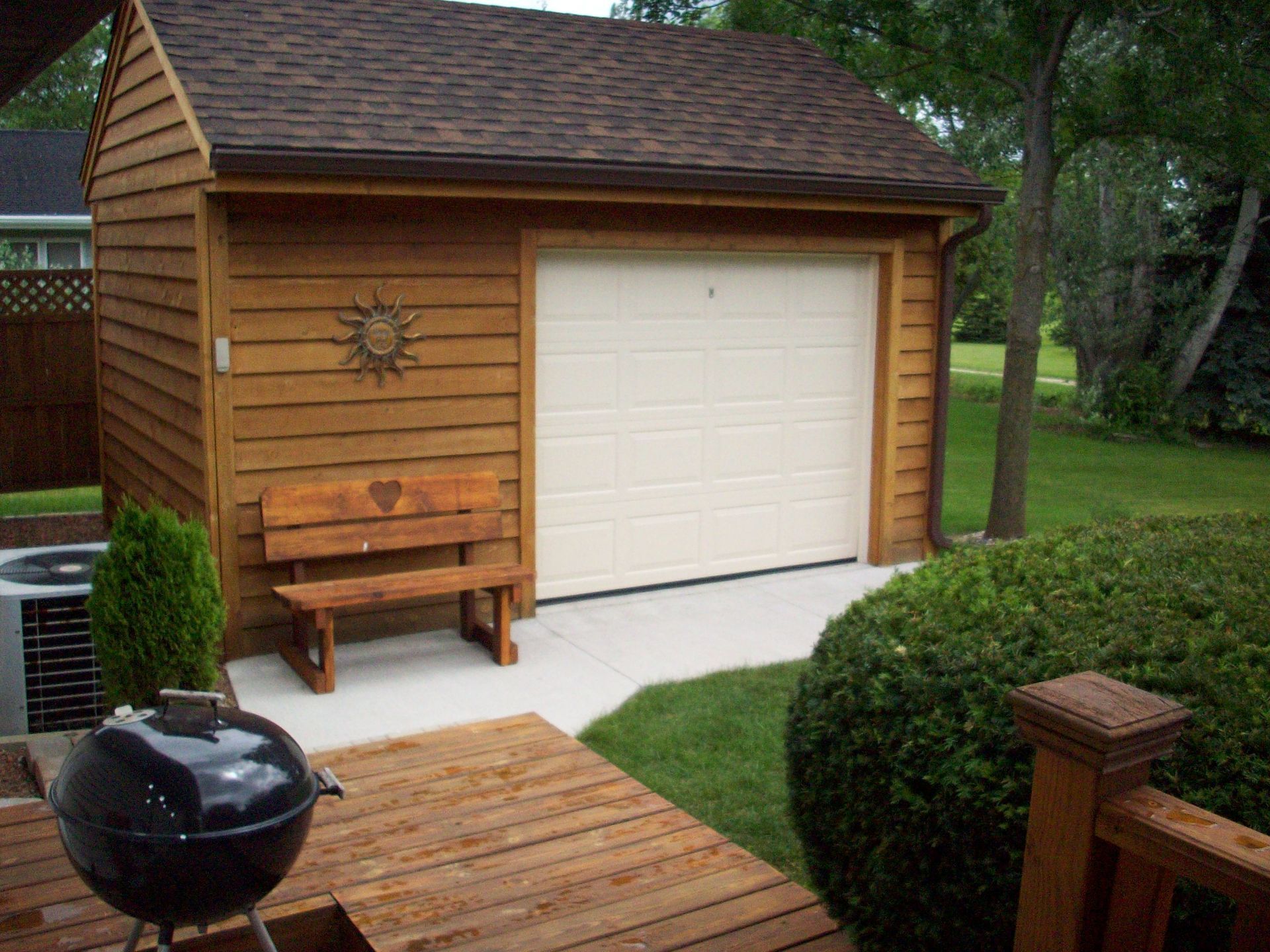 Wooden backyard shed with white garage door, grill, bench, and deck beside shrubs and lawn