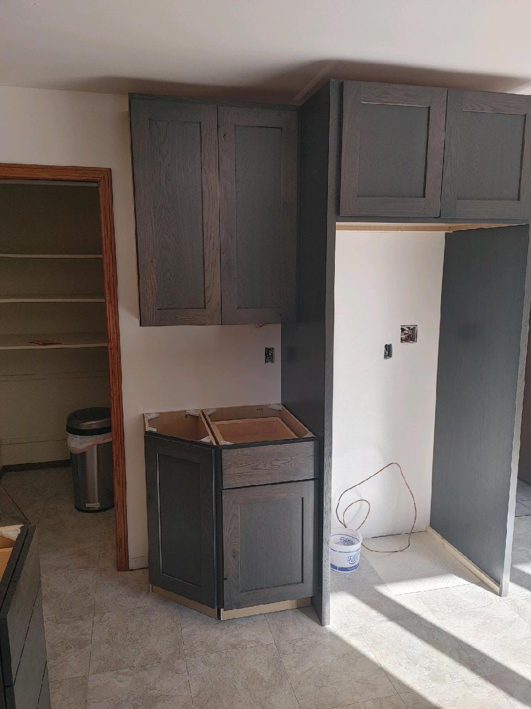 Empty kitchen with unfinished gray cabinets, white walls, and sunlight on the floor.