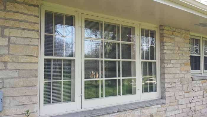 White-framed window set in a stone house wall with blinds and reflections of trees outside.