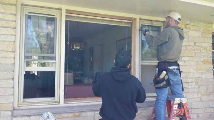 Two workers installing a large window in a stone house exterior.