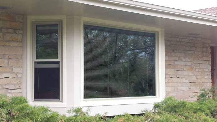 Stone house exterior with a large white-framed window and blinds, plus a narrow side window and shrubs below