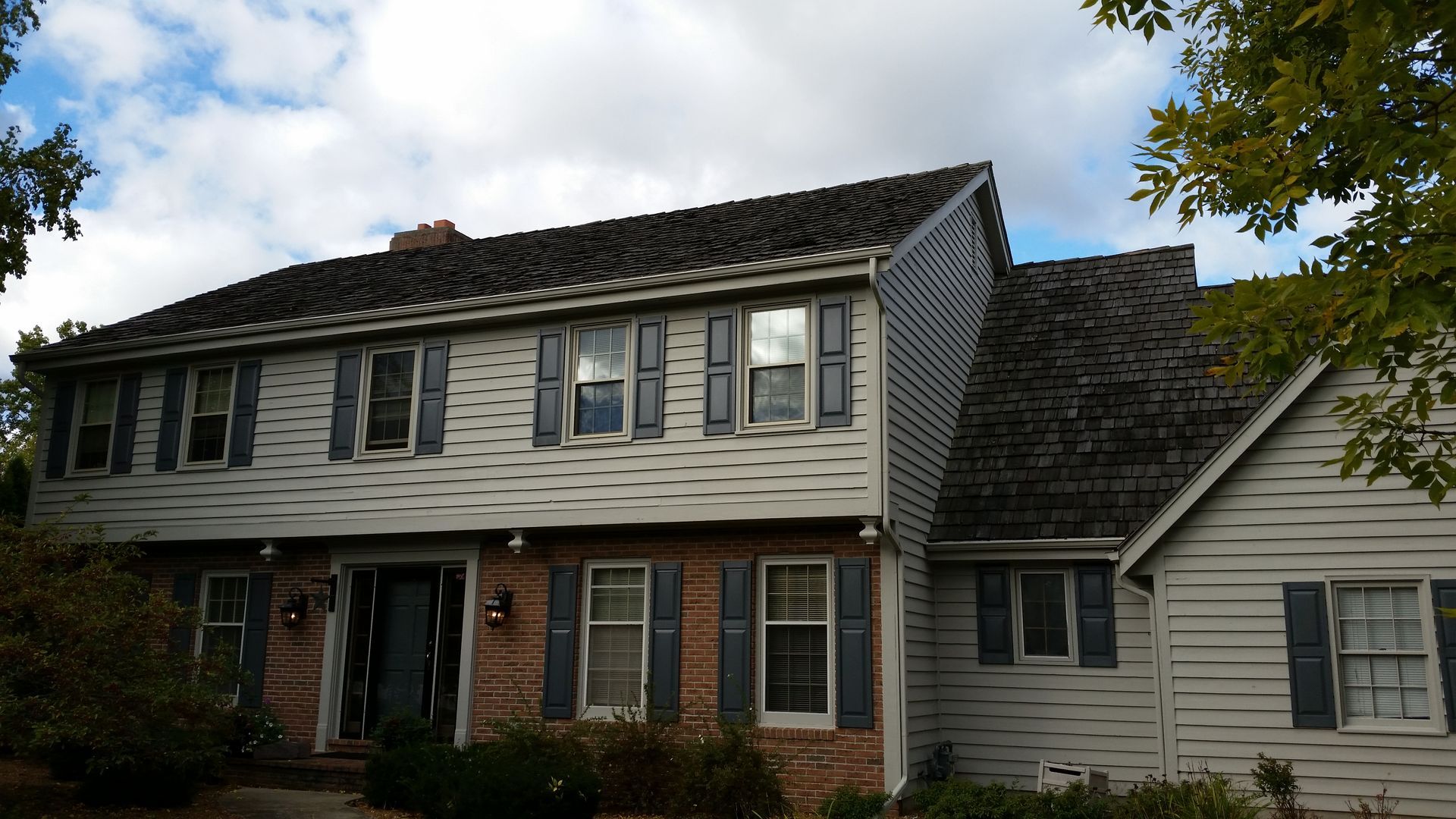 Two-story gray house with dark shingled roof, white trim, and front porch under a partly cloudy sky