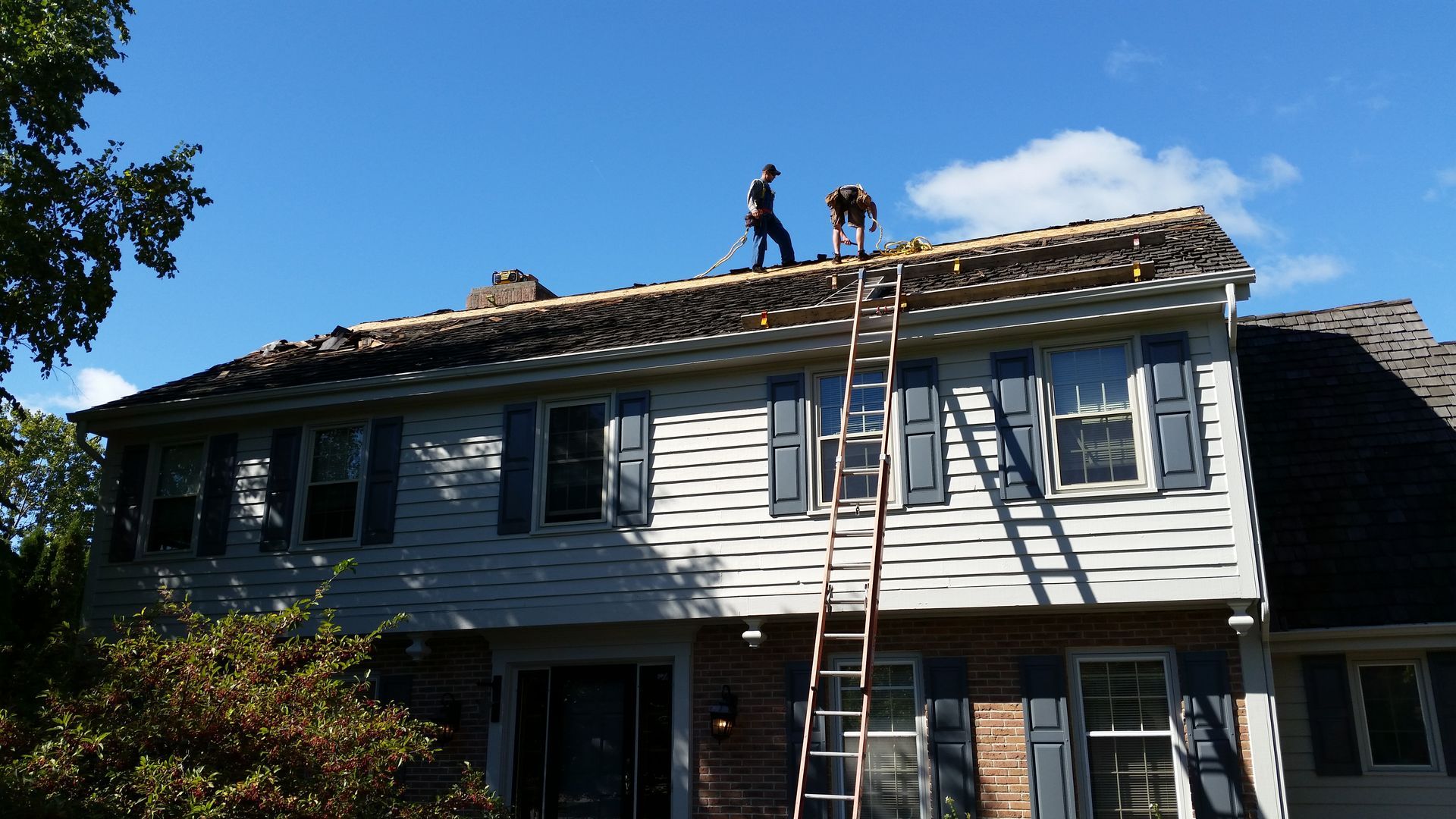 Workers on a house roof during repair, with a ladder leaning against the siding.
