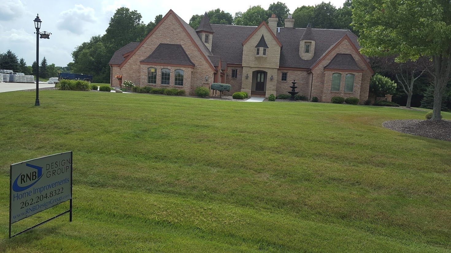 Large stone house with a steep roof on a grassy hill, with a sign in the foreground and trees behind.