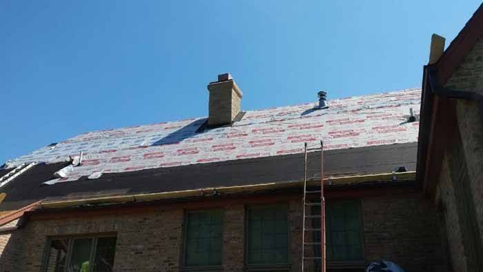 House roof under repair with white underlayment, a ladder, and blue sky above.