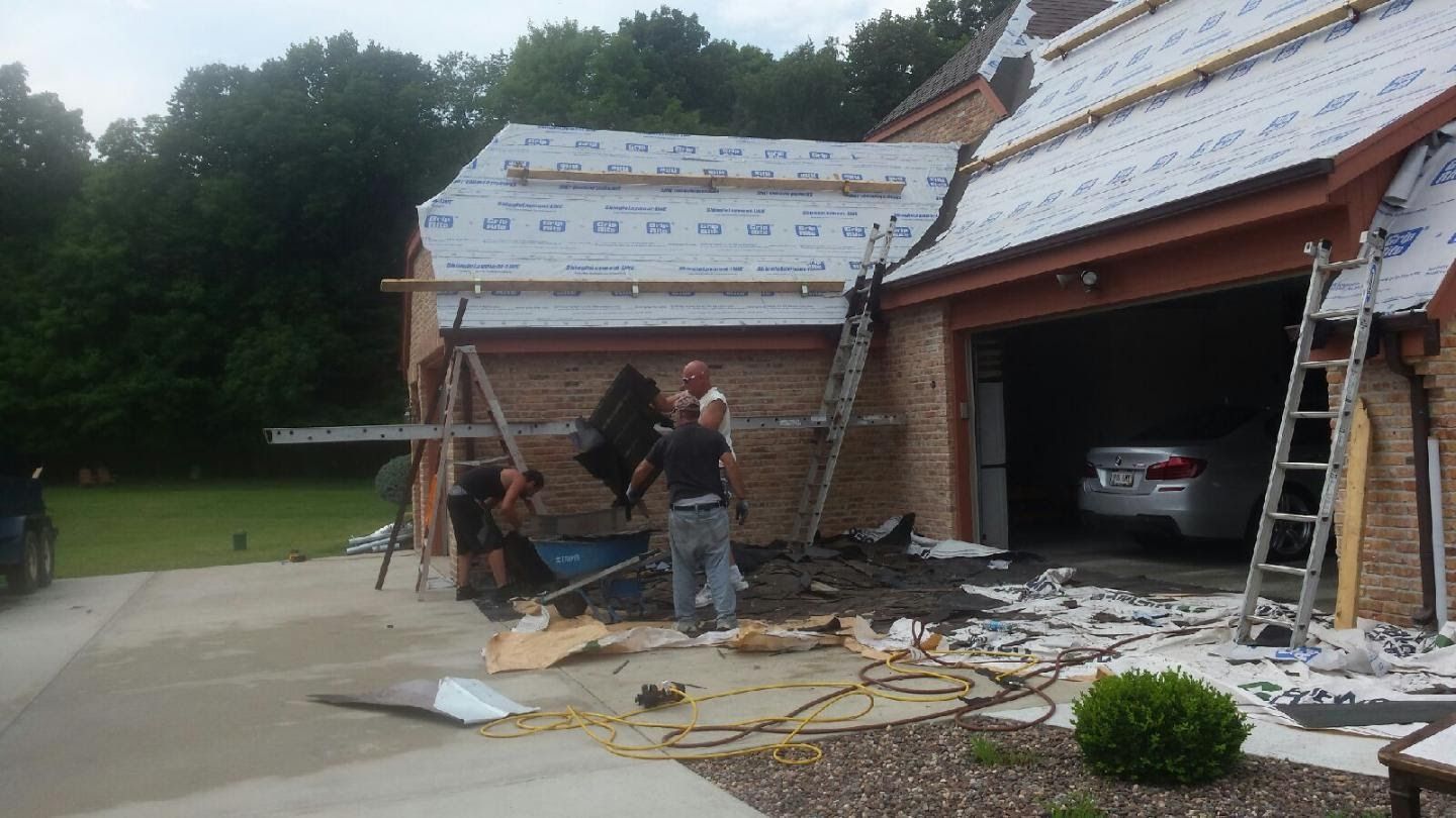 Workers repairing a brick house roof and garage after storm damage, with debris and ladders nearby